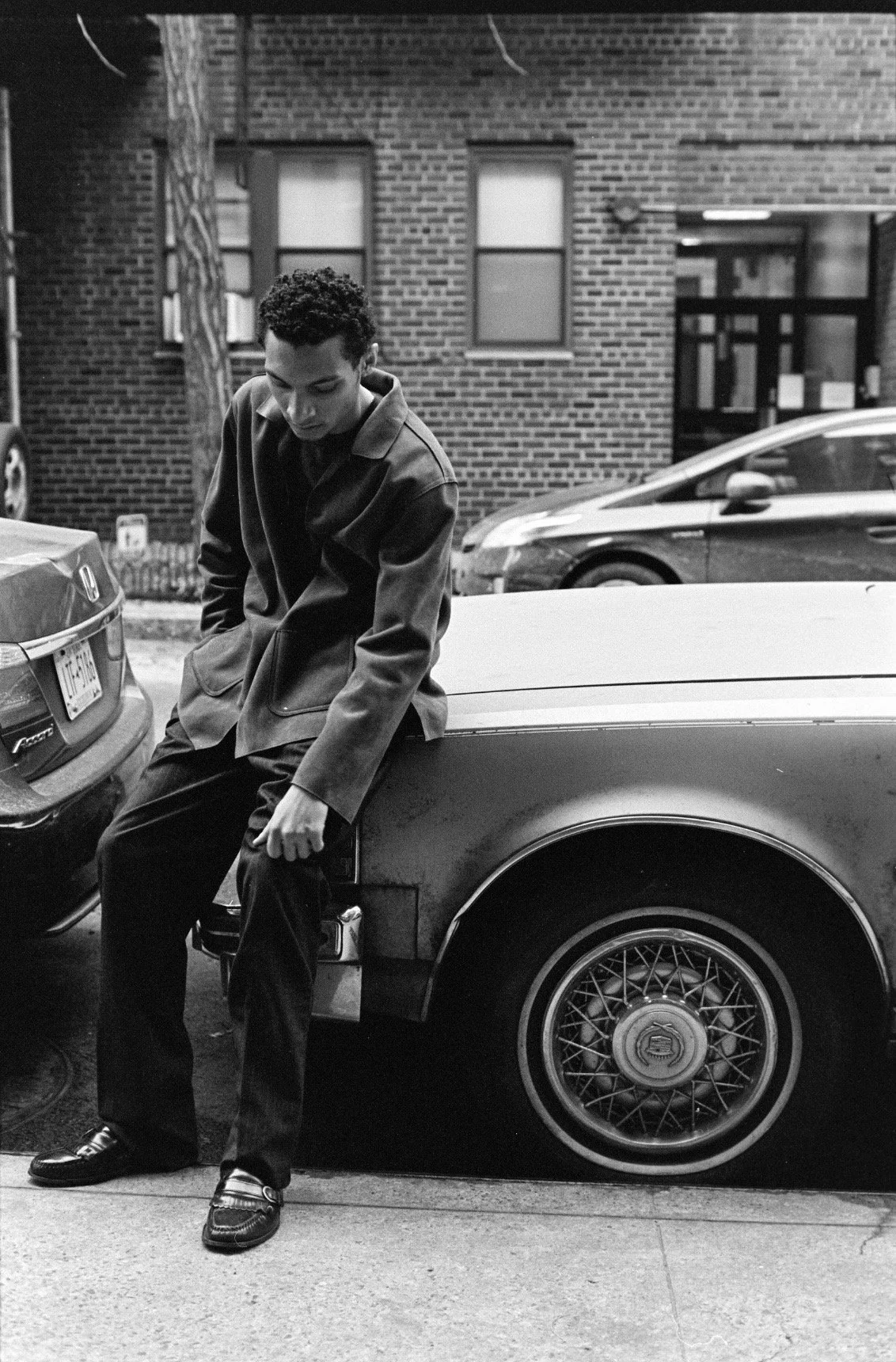 A man with curly hair wearing a dark jacket and pants, sitting on the rear fender of a car parked on the street in an urban setting. Multiple cars and a brick building are in the background.