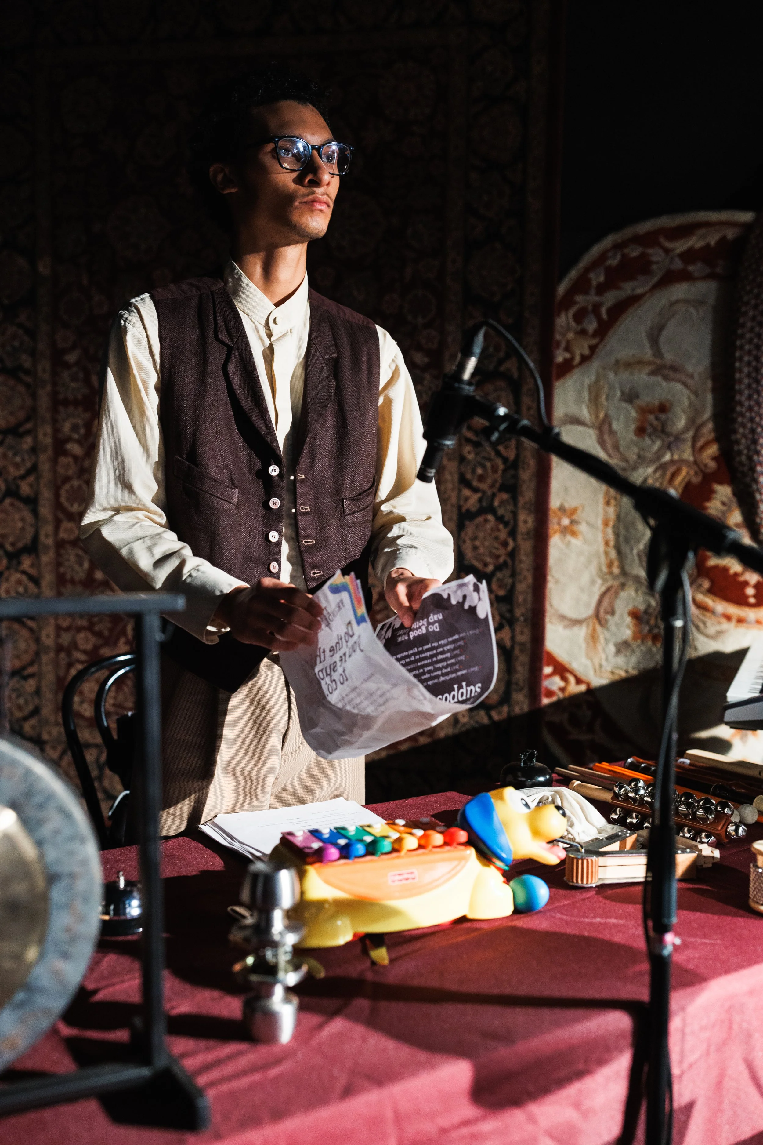 A young man with glasses, wearing a white shirt and dark vest, stands behind a table with a paper in his hand. The table has various musical and theatrical objects, including a colorful puppet and a xylophone, with a microphone set up in front of him