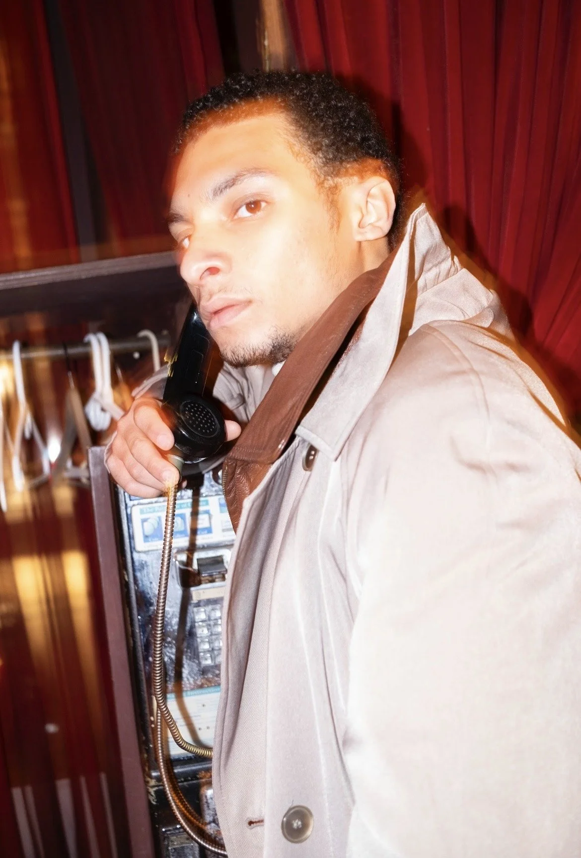 A young man with short curly hair and light skin holding a phone receiver to his ear, standing in front of a payphone in a phone booth with wood paneling.