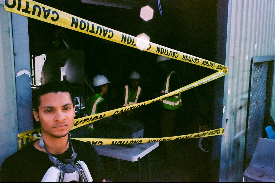 A young man standing in front of a construction or industrial area, with three workers in hard hats and safety vests inside a cordoned-off zone. Yellow caution tape blocks the entrance to the area.