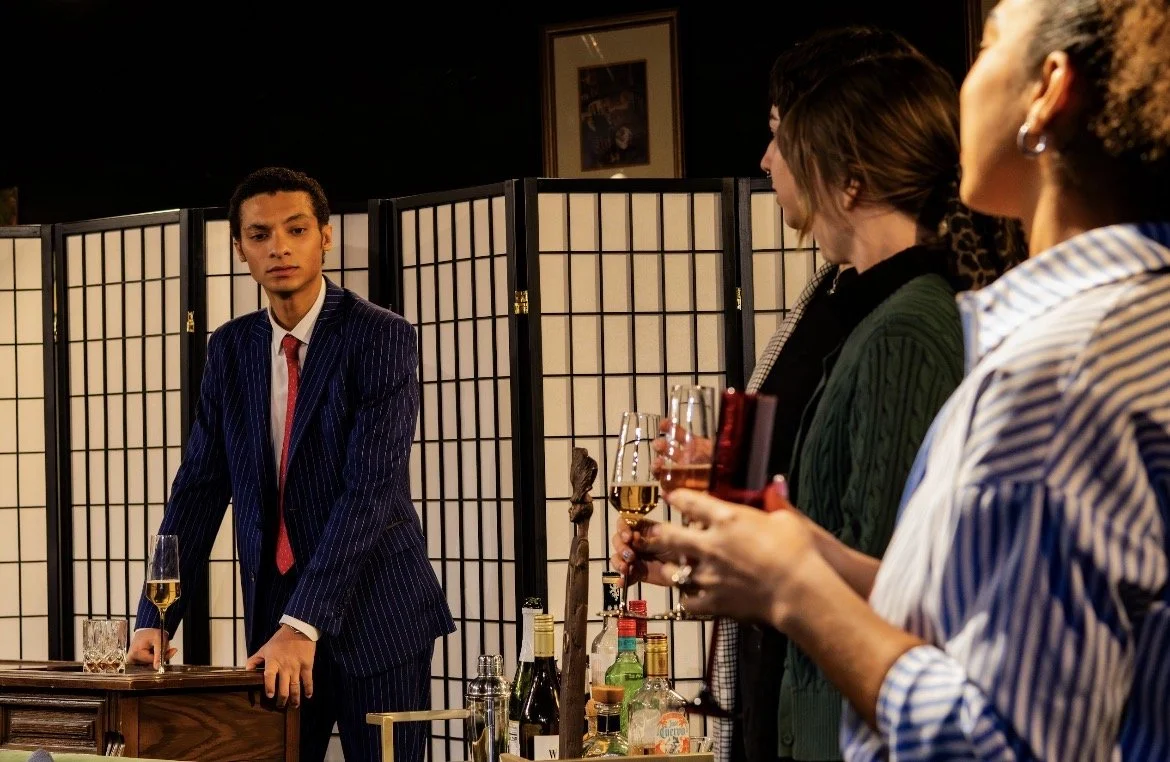 A man in a dark pinstripe suit and red tie appears apprehensive as he stands behind a bar table, while three women hold wine glasses and face him in a dimly lit room with a folding screen and framed picture in the background.