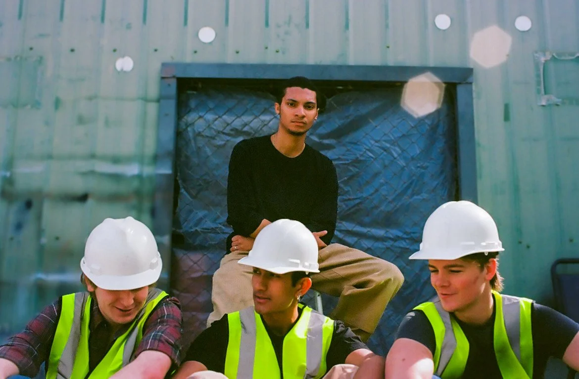 Group of construction workers wearing white helmets and high-visibility vests, with a young man sitting on a platform wall behind them, inside a building with green metal walls.