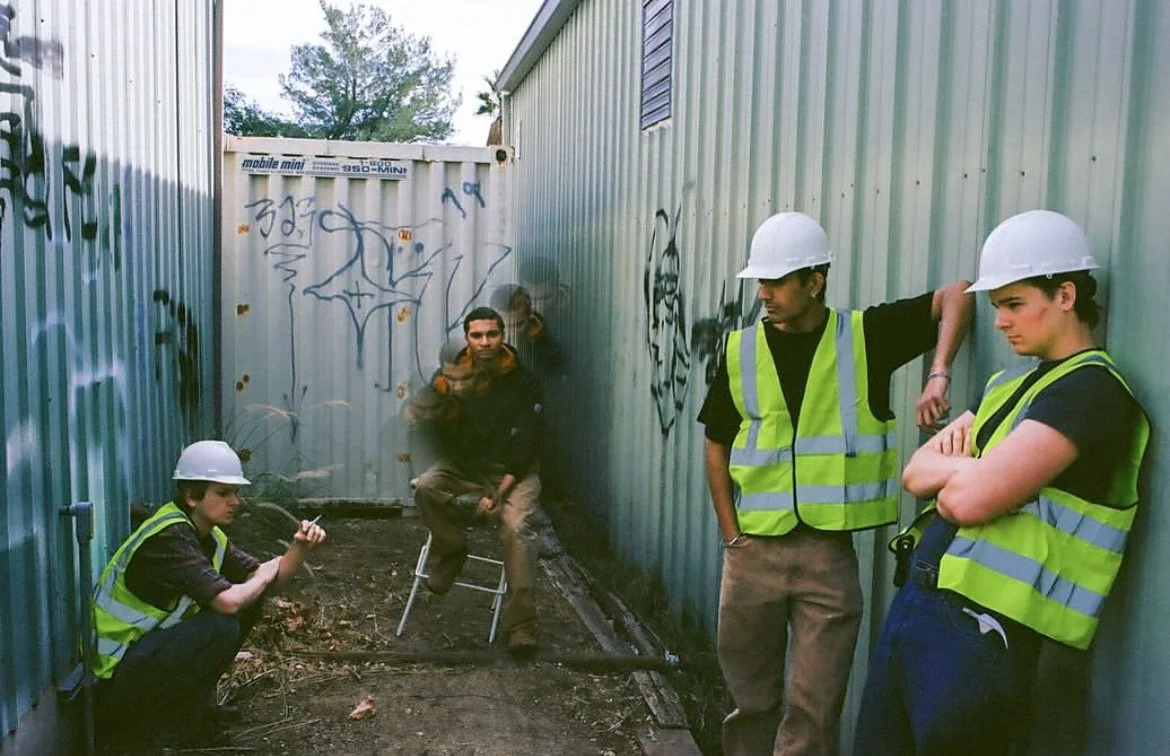 Four construction workers wearing white safety helmets and reflective vests in a narrow alleyway with graffiti-covered corrugated metal walls. One worker is sitting on a small ladder, one is squatting and holding a phone, and two are standing and lea