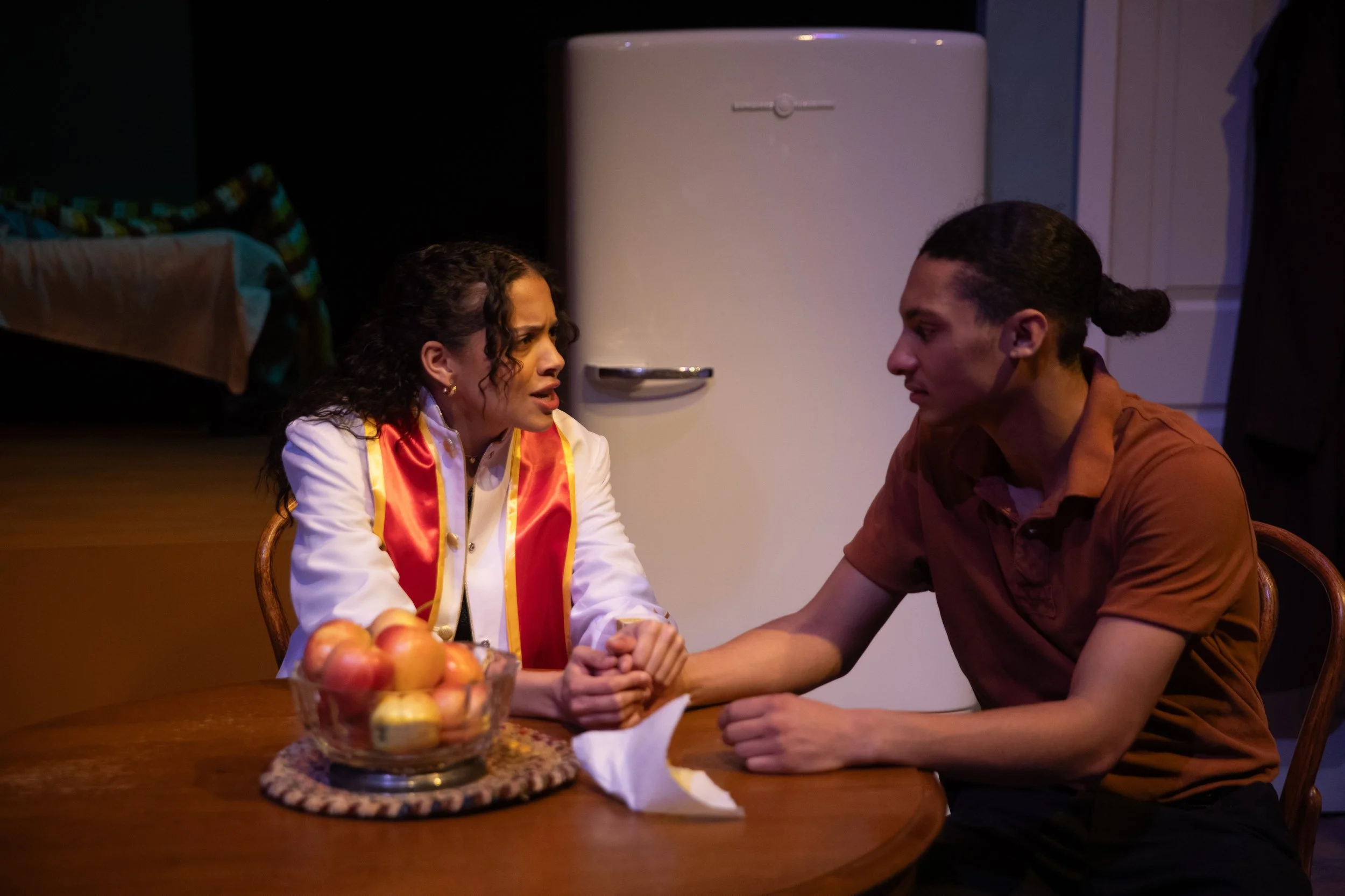 A woman and a man sit at a wooden dining table holding hands and looking at each other. The woman appears emotional, and there is a bowl of apples on the table behind them. In the background, a white refrigerator and a bed are visible in a domestic s