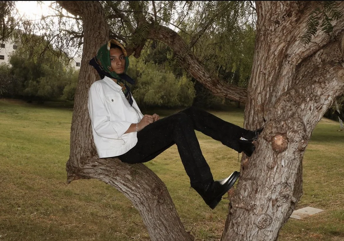 A person sitting on a tree branch in a park, wearing a white jacket, black pants, boots, and a headscarf, with a serious expression.