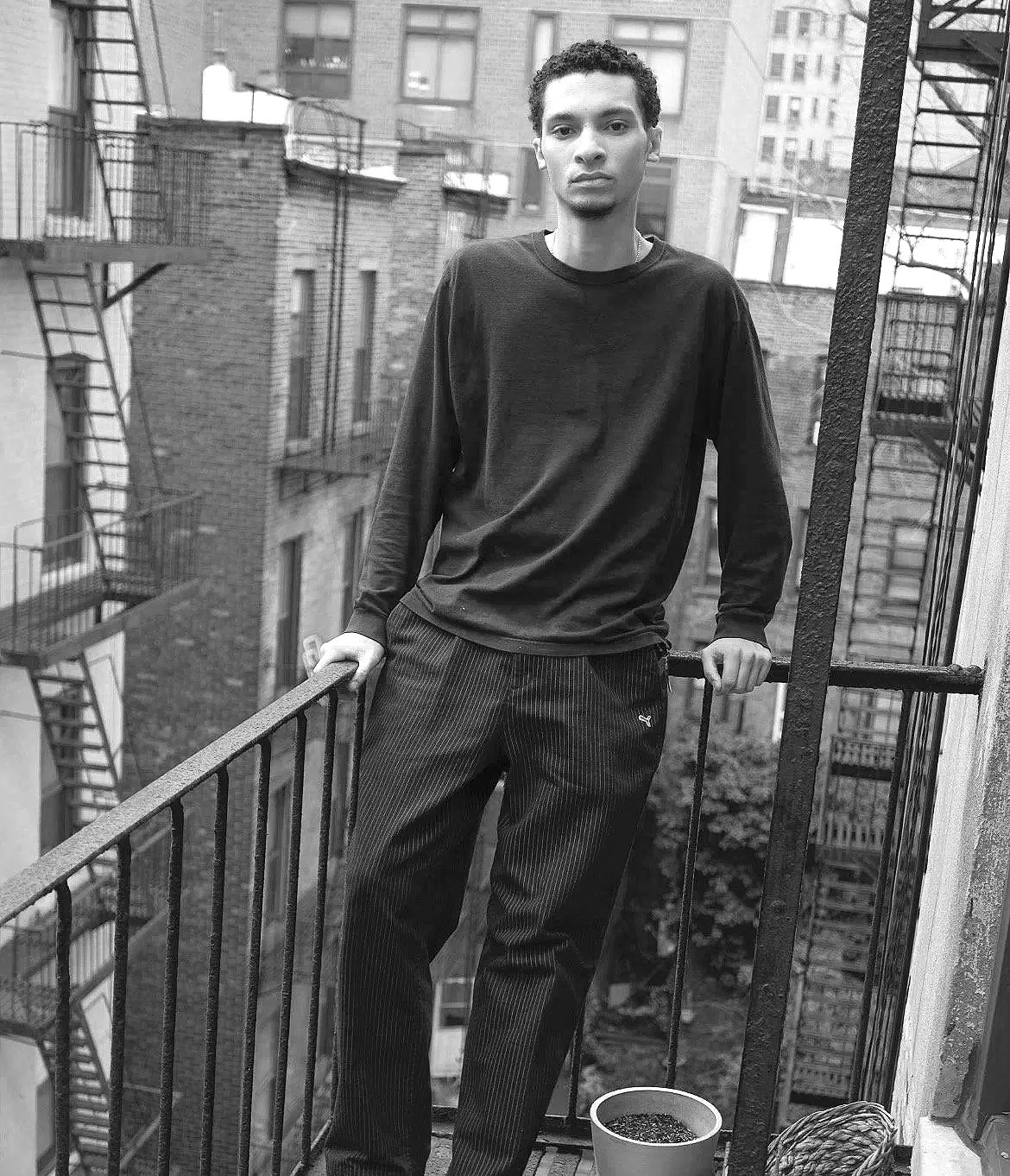 A young man standing on a small balcony in an urban neighborhood, wearing a black long sleeve shirt and striped pants, with buildings and fire escapes in the background, and a bucket of water or soil on the balcony.