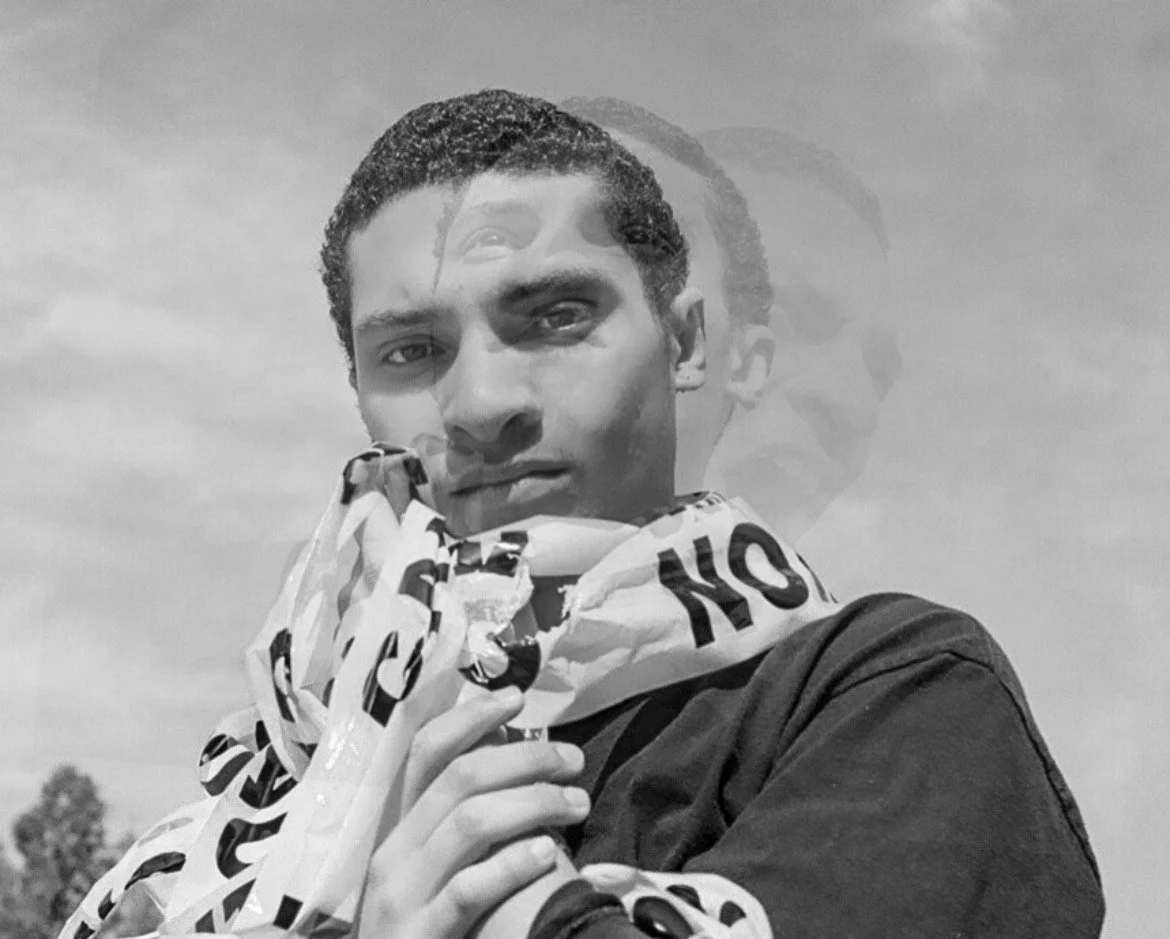 Black and white photo of a young man with short curly hair, looking directly at the camera, with a faint, semi-transparent second image of himself in the background, outdoors with a cloudy sky and trees in the distance.
