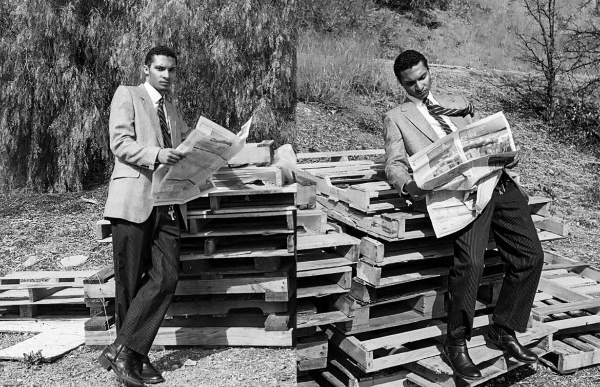 Two young men in suits leaning against wooden pallets while reading newspapers outdoors in black and white.