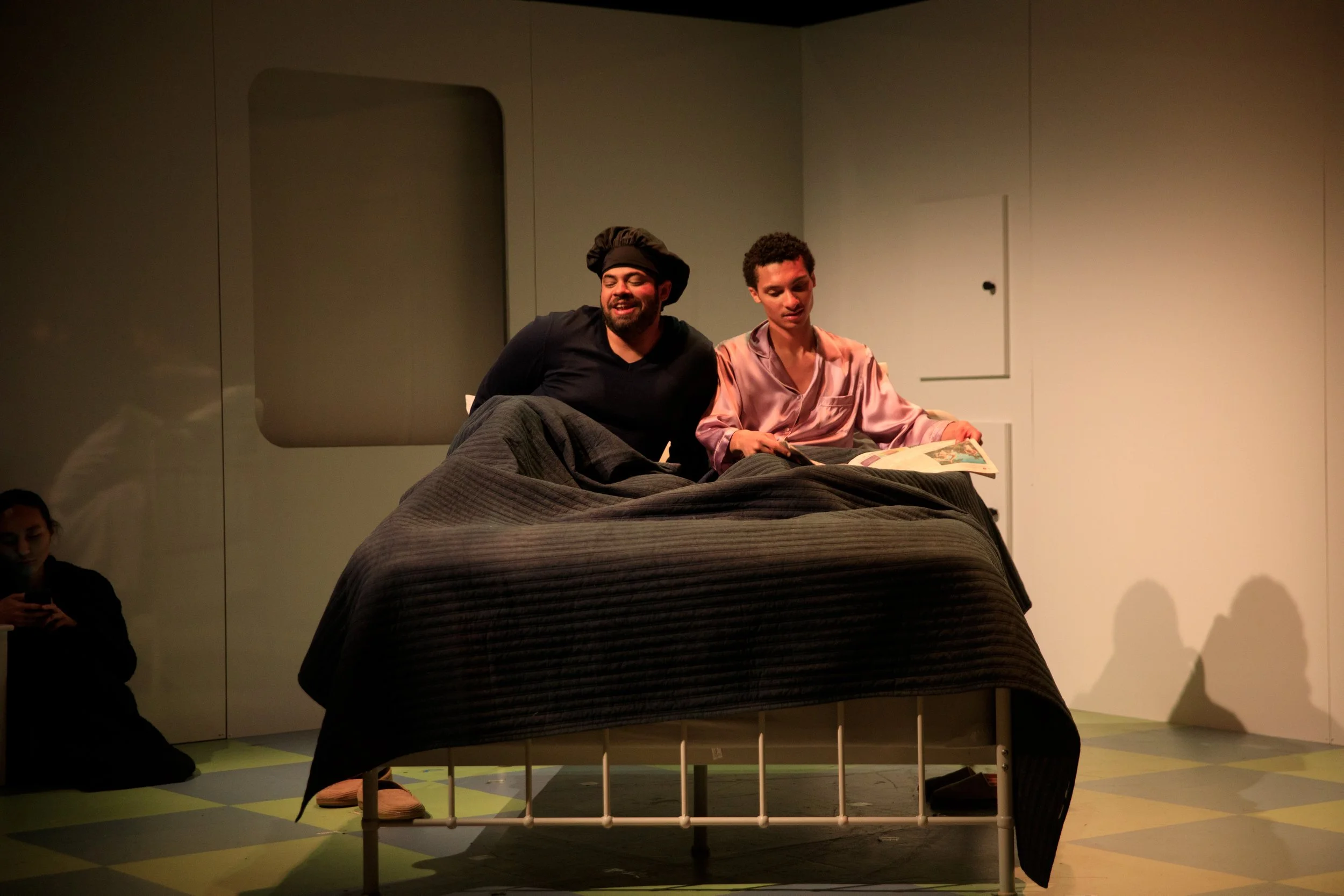 Two actors sit on a bed in a minimalistic room, one wearing a pink satin robe reading a magazine, the other smiling and wearing a black shirt and hat.