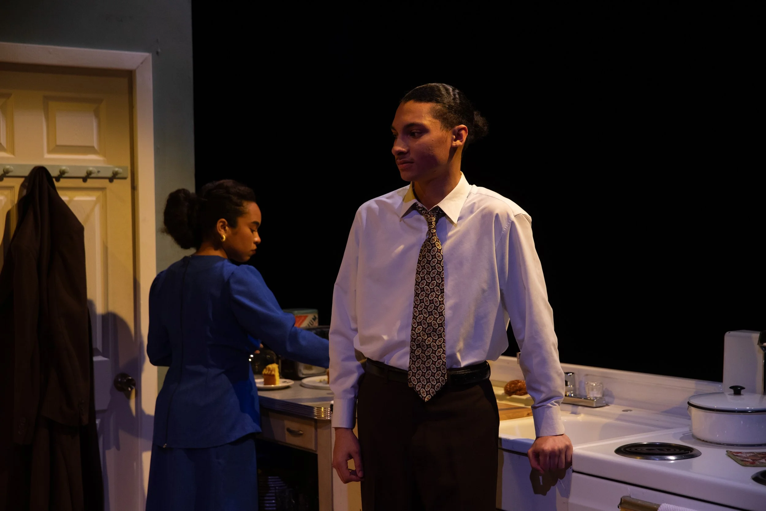 Two actors on stage, a woman in a blue dress preparing food at a kitchen counter, and a man in a white shirt and patterned tie standing nearby, with a black background.