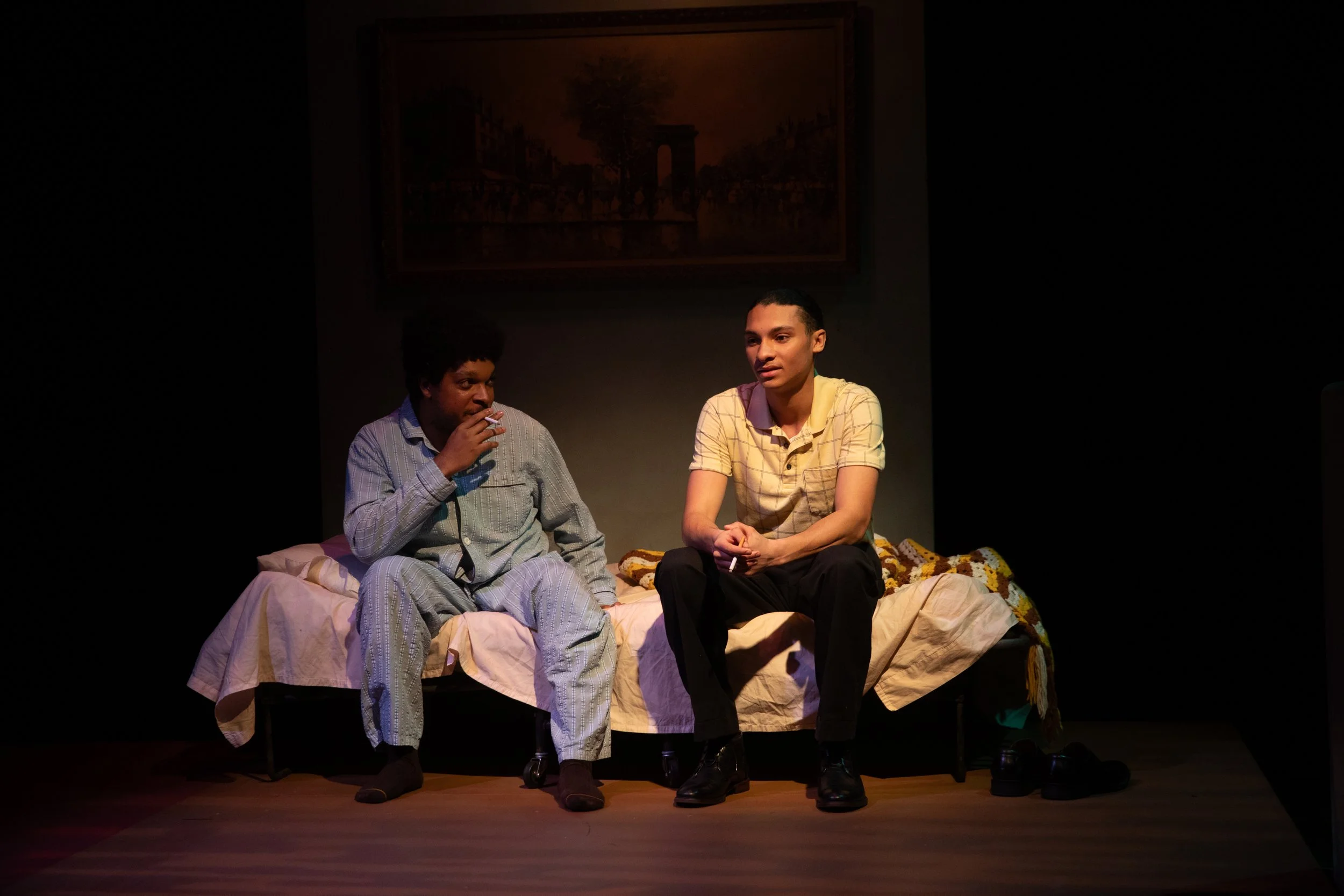 Two men sitting on a bed in a dimly lit room, engaged in conversation. One is wearing pajamas and smoking, the other is dressed in casual clothes. A painting hangs on the wall behind them.