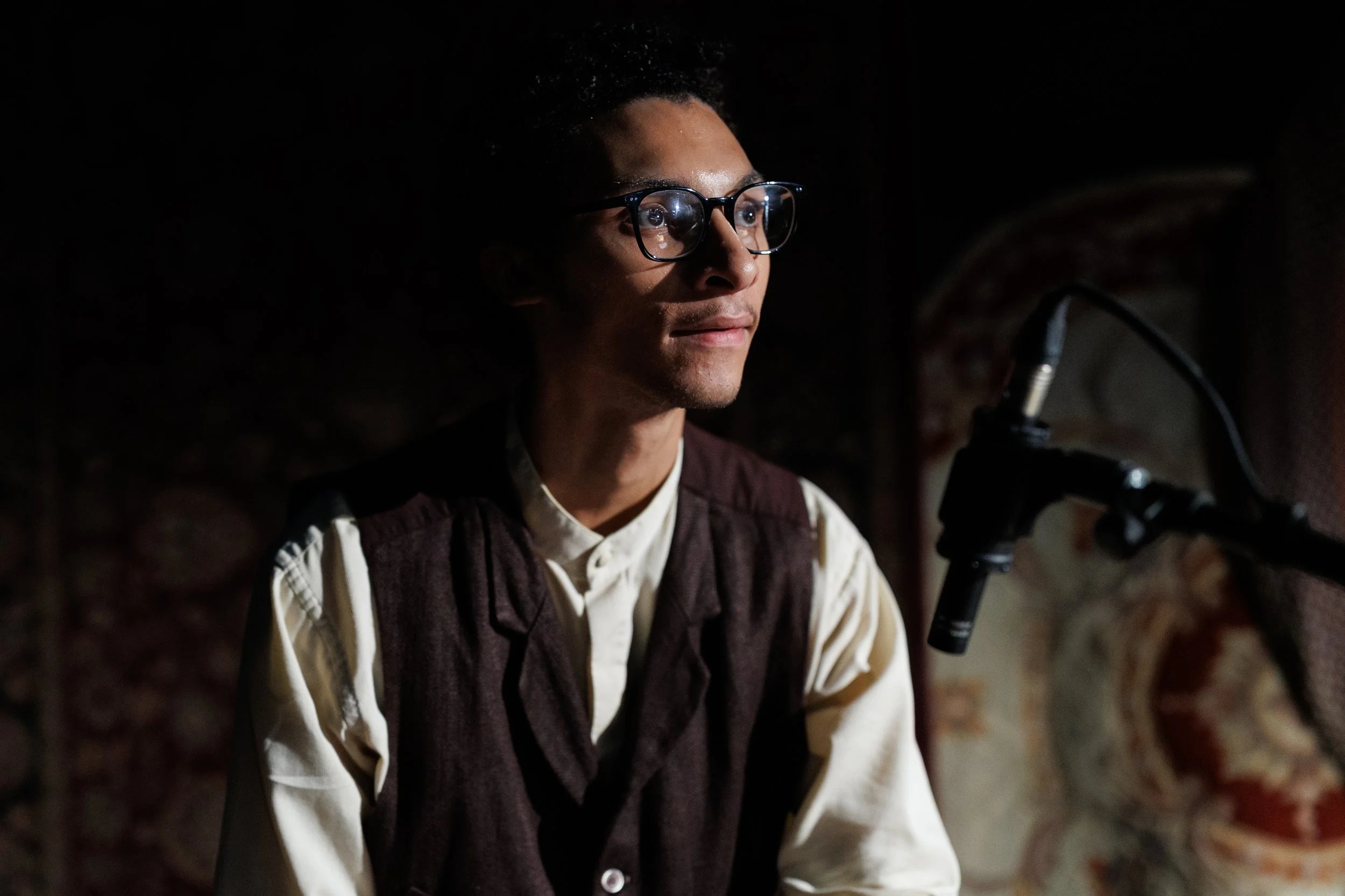 A man wearing glasses and a vintage vest sits in front of a microphone in a dimly lit room, with tapestries on the wall behind him.