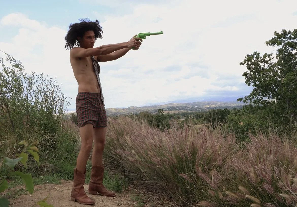 A person standing outdoors aiming a green toy gun, wearing shorts and boots, with a natural landscape and cloudy sky in the background.