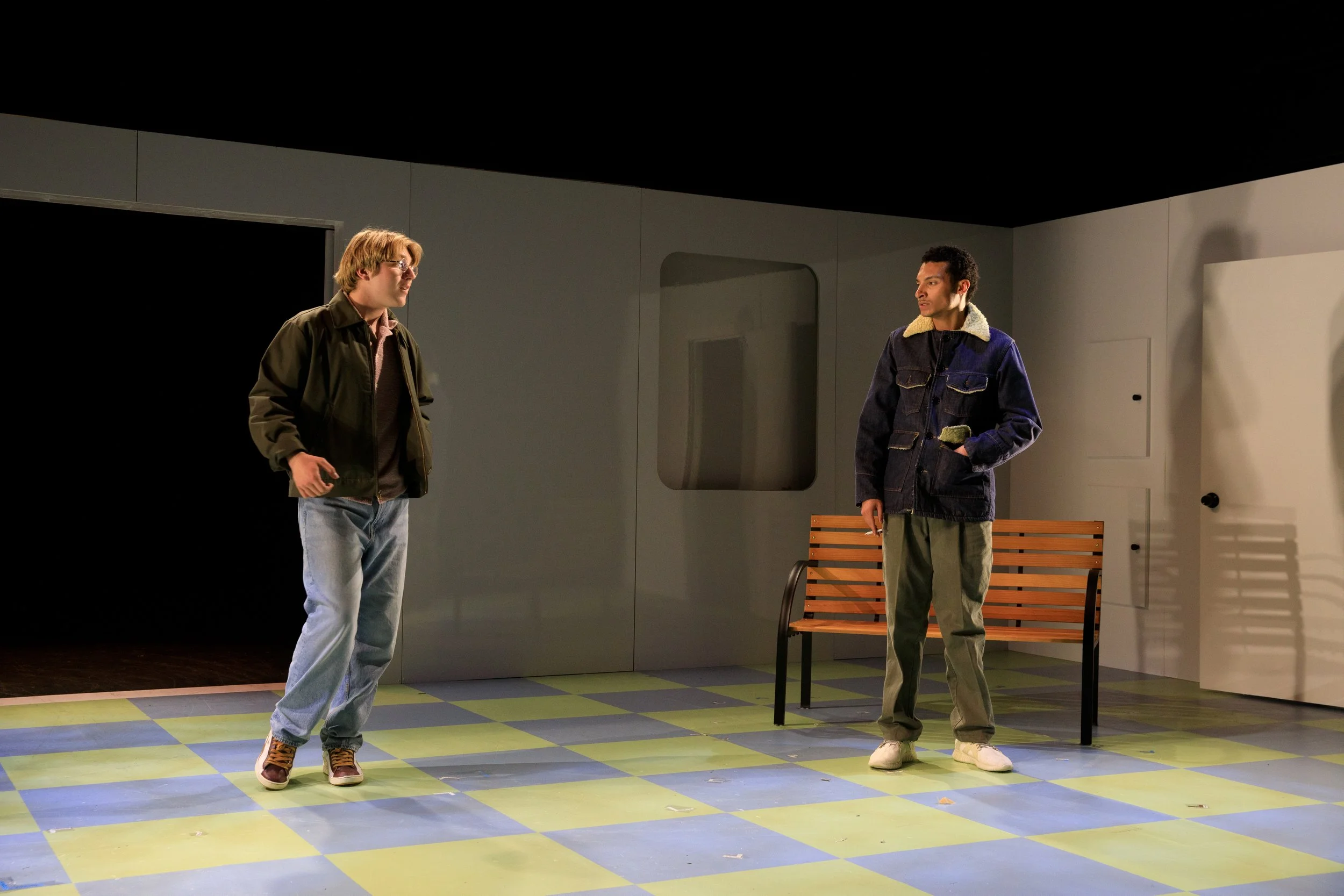Two men stand on a stage with a bench behind them, engaged in conversation. One has blonde hair, glasses, a brown jacket, and jeans. The other has dark hair, a dark jacket with a shearling collar, and green pants.