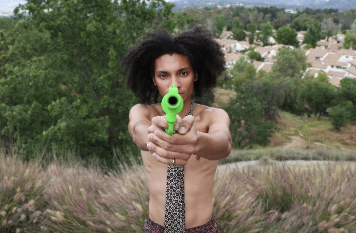 A shirtless woman with curly hair holding a bright green toy gun pointed at the camera in an outdoor setting with greenery and houses in the background.