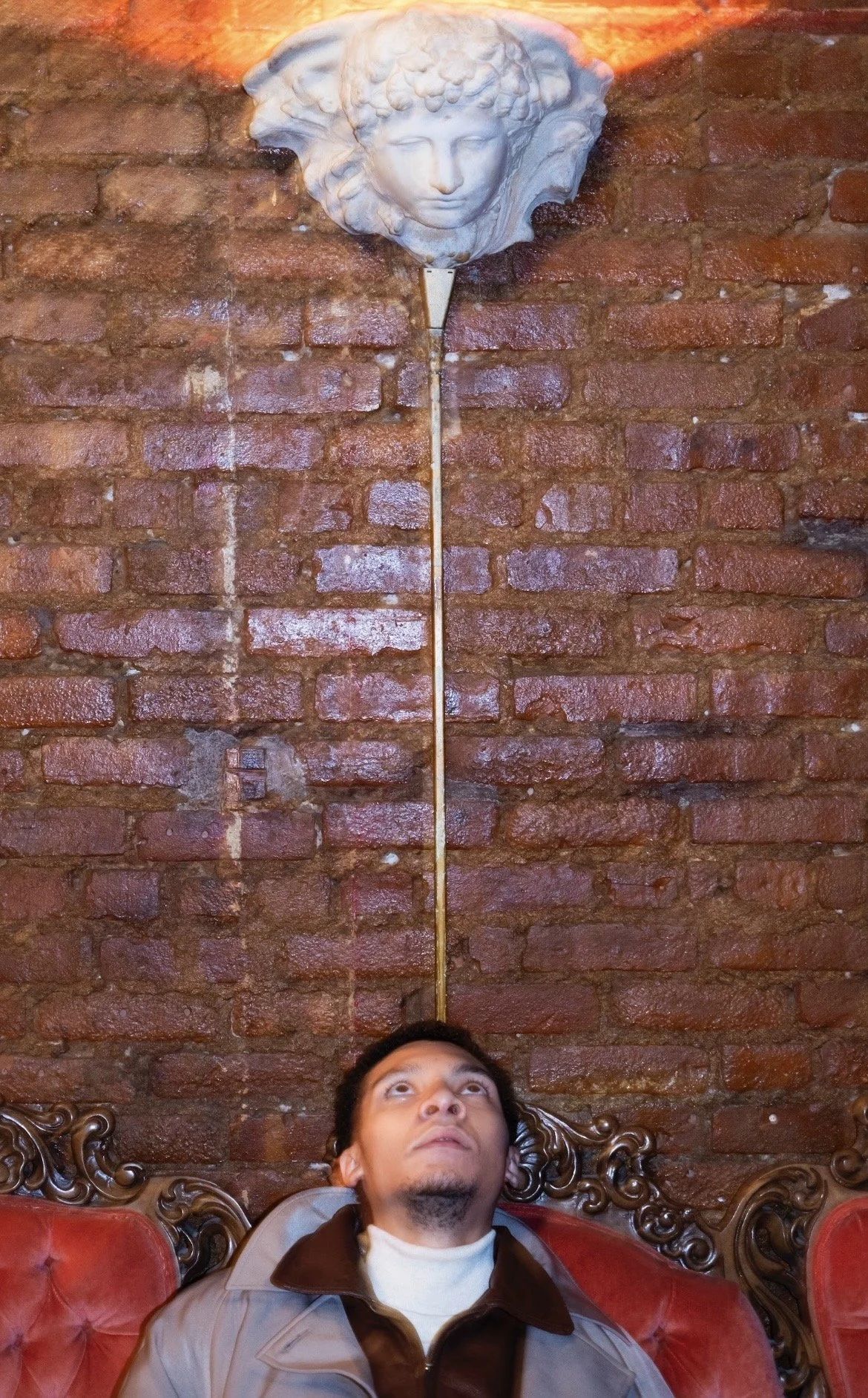 A man looking up while sitting on a red, vintage-style sofa with ornate wooden details. Behind him, there is an exposed brick wall with a decorative wall sculpture of a woman's face with flowing hair, illuminated from above.