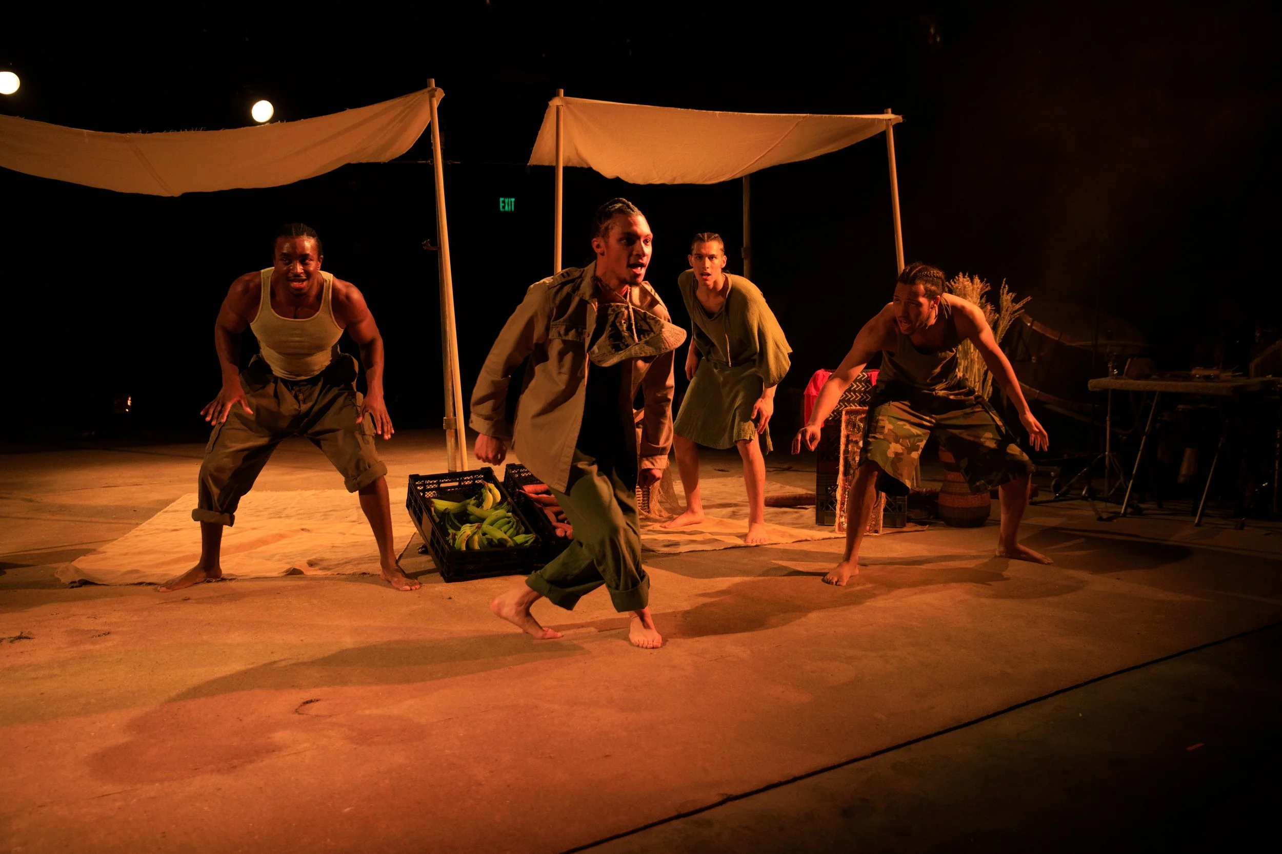A theatrical performance on stage featuring five actors in casual costumes, positioned in dynamic poses under warm lighting, with a backdrop of simple props and a canopy overhead.
