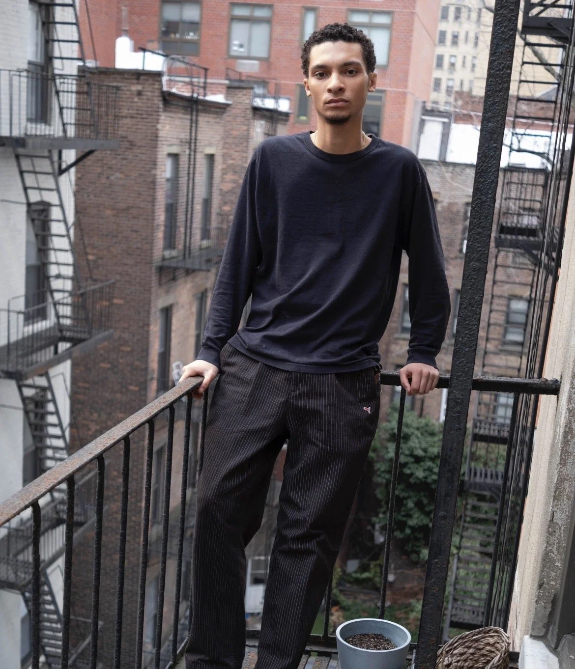 A young man with curly hair and a serious expression standing on a small balcony in an urban setting, wearing a black long-sleeve shirt and dark pinstriped pants, holding onto the railing with both hands, with brick apartment buildings and fire escap