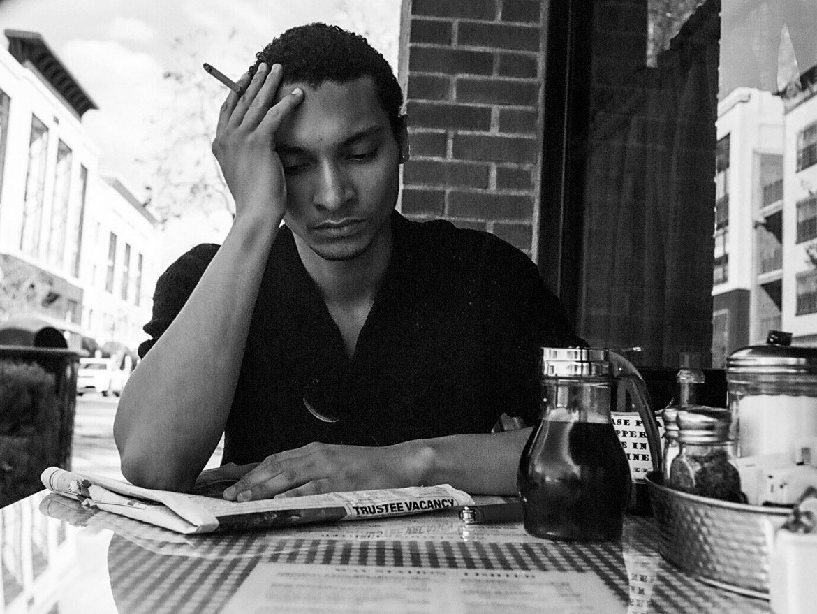 A young man sitting at a table in a cafe, resting his head on his hand, reading a newspaper, with condiments and a pitcher of dark liquid on the table, on a city street.
