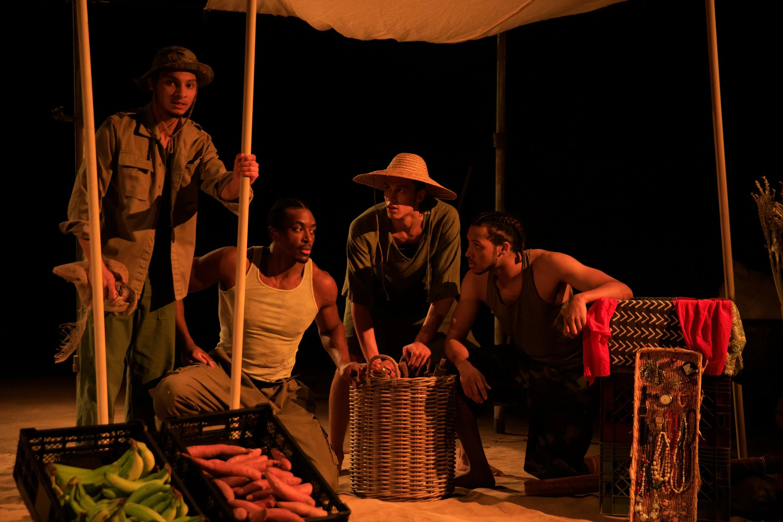 Five actors on stage in a theater performance, dressed in costume, some sitting and some standing, with various props including baskets of sweet potatoes and bananas, a crate with jewelry, and a small table with a patterned cloth, under a canopy at n