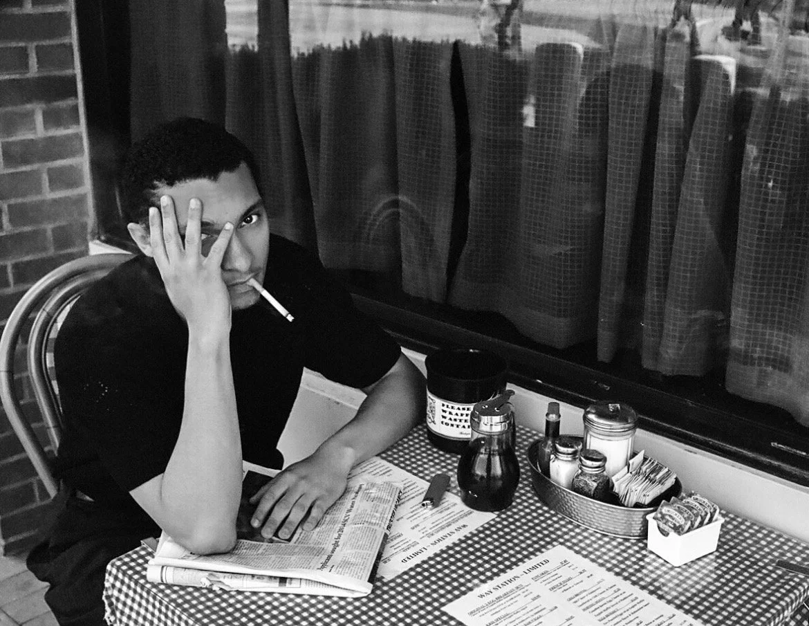 A young man sitting at a table in a restaurant, holding his face with one hand, smoking a cigarette, and looking at the camera. The table has a checkered tablecloth, condiments, and a newspaper.