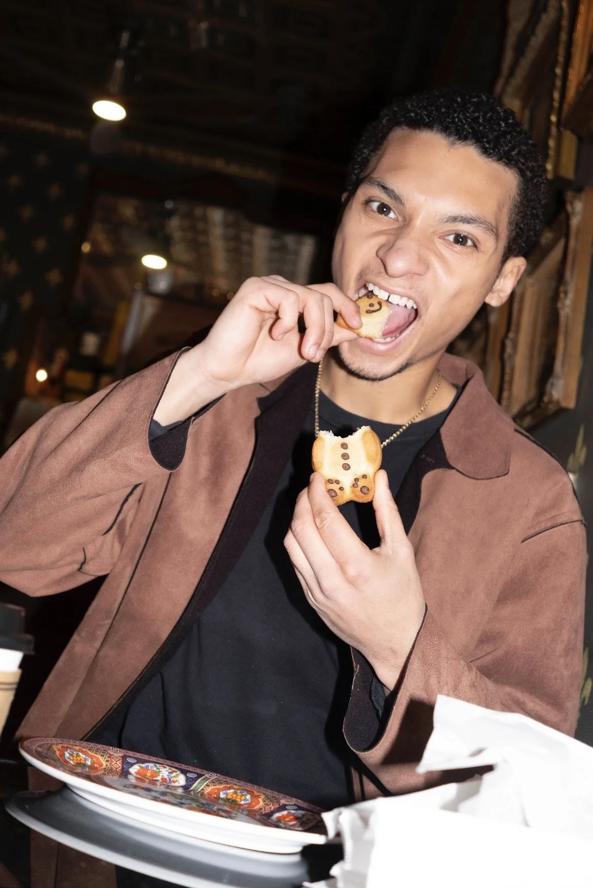 A young man with dark curly hair and a brown jacket is eating decorated cookies shaped like bears in a restaurant or café.