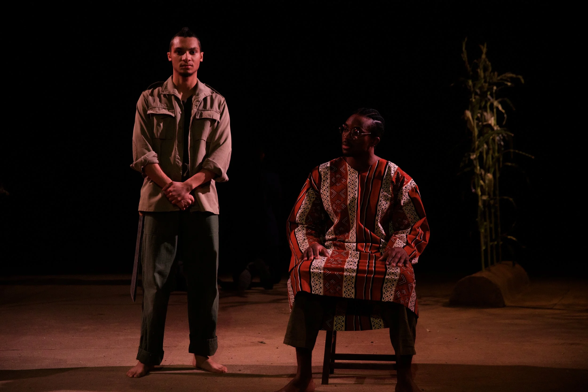 Two men on stage, one standing and one seated, under dark lighting with a plant in the background.