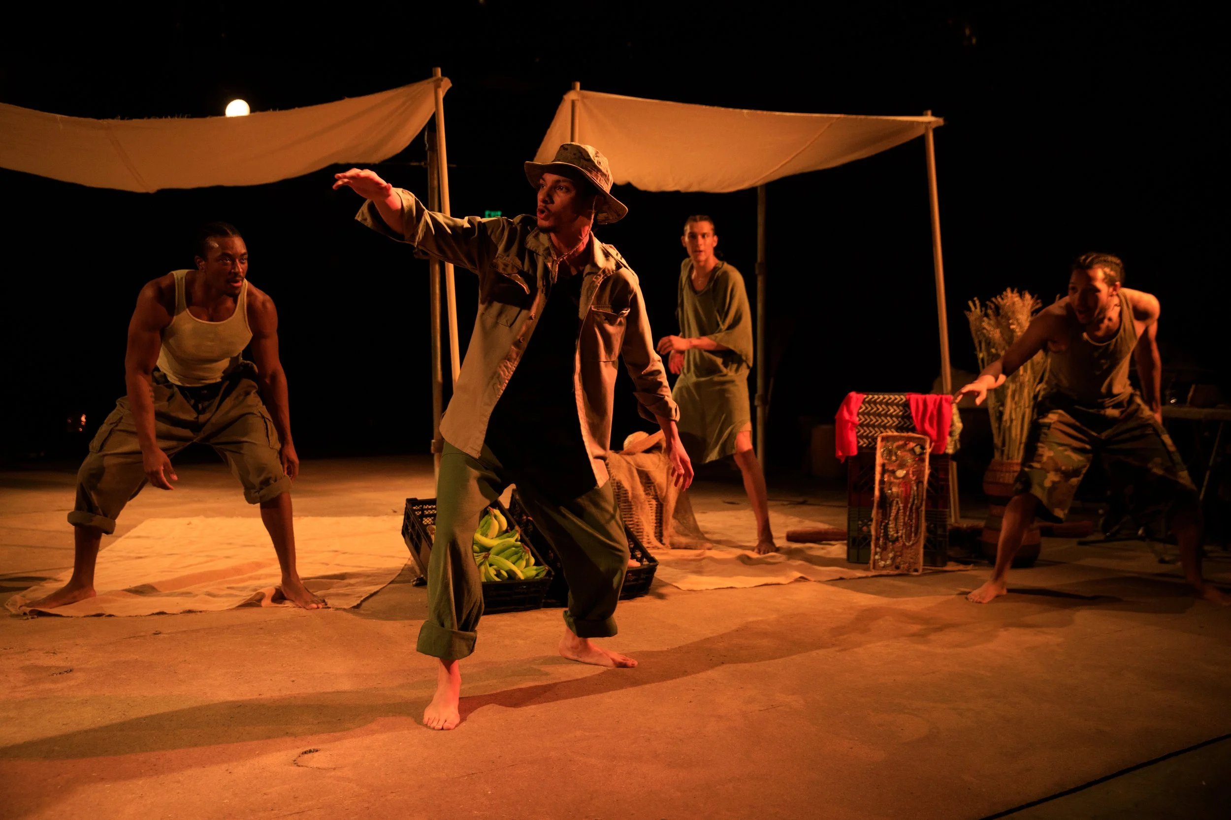 Four performers in costume on stage during a theatrical performance, with props and fabric drapes in the background, under warm lighting.