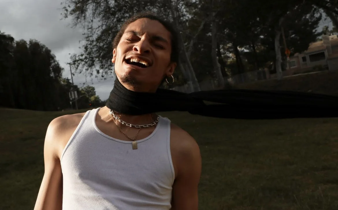 Person singing or shouting outdoors, wearing a white tank top, silver necklaces, and hoop earrings, with trees and a house in the background.