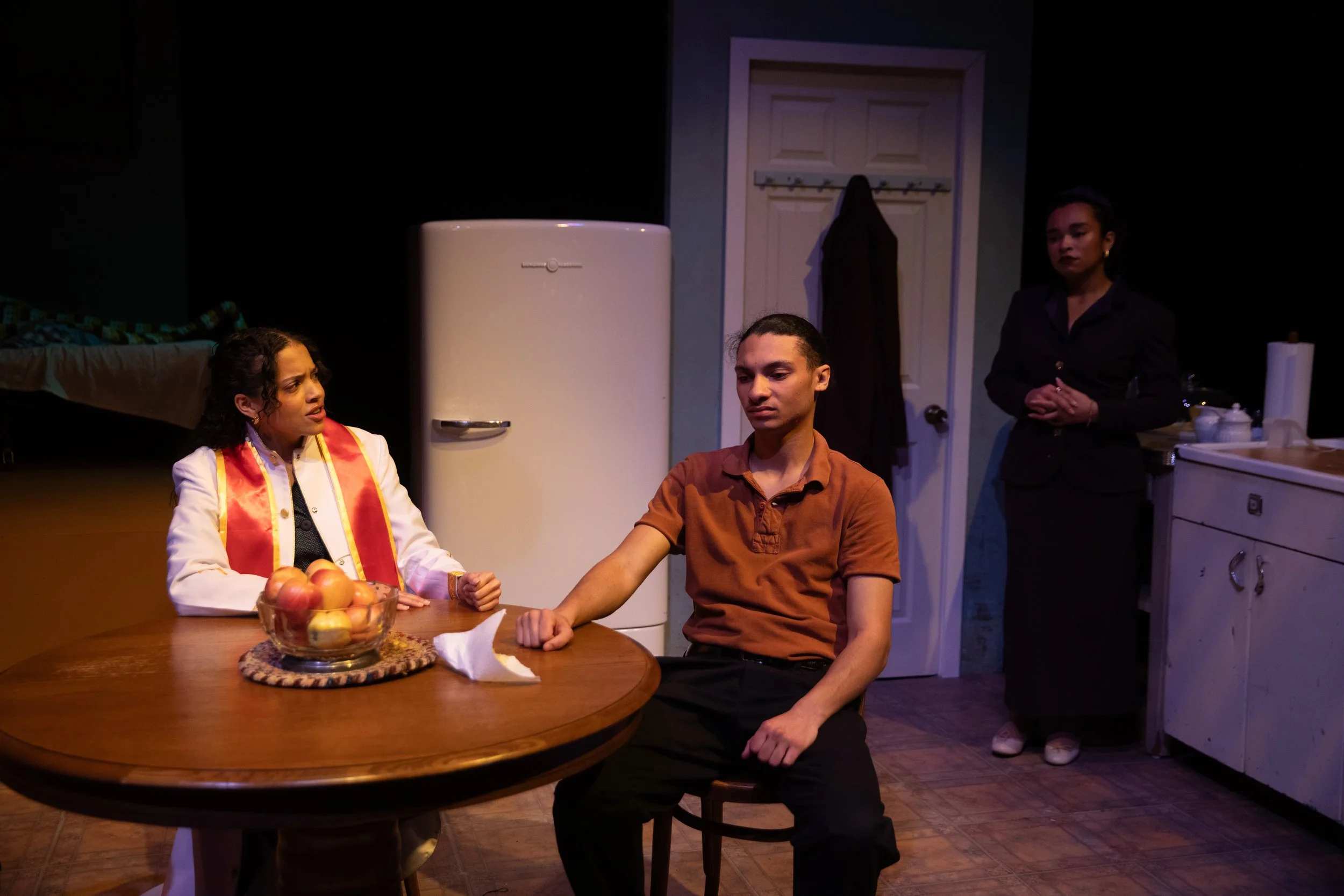 Three women in a kitchen scene with a young man sitting at a round table. One woman is sitting and talking, another is standing behind the table, and the third woman is standing near the counter. A bowl of apples and a white paper towel are on the ta