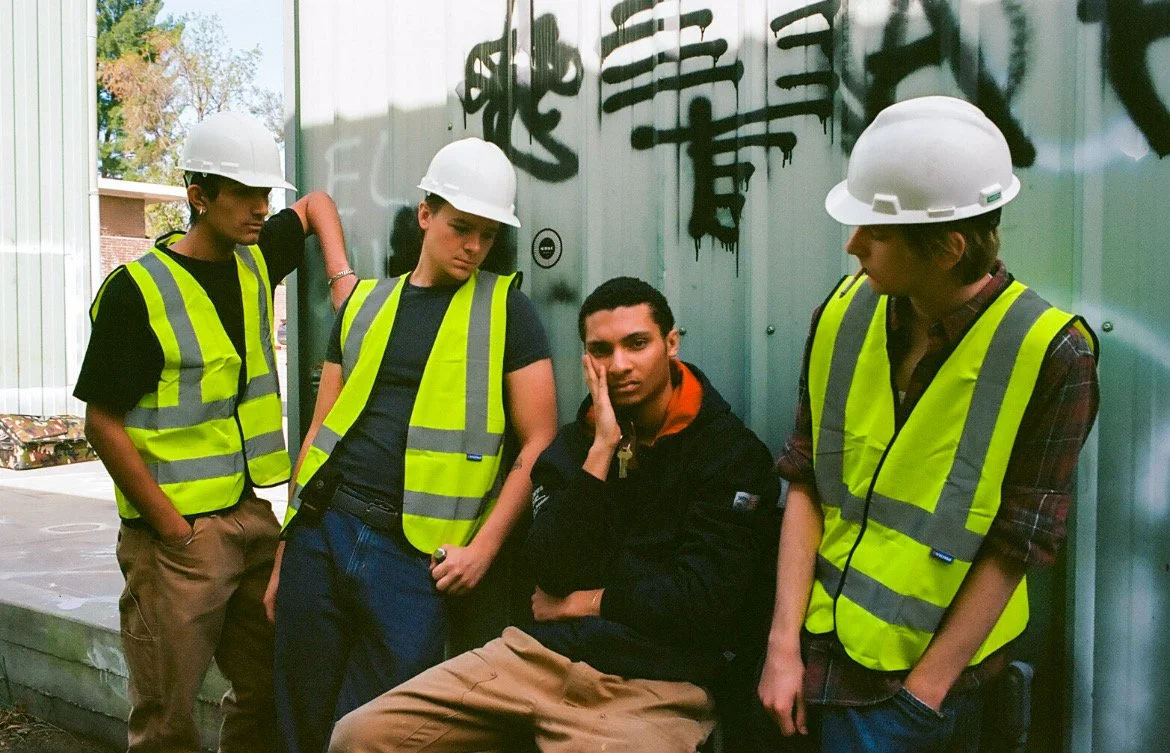 Four young people wearing safety vests and hard hats standing and sitting near graffiti on a metal wall.