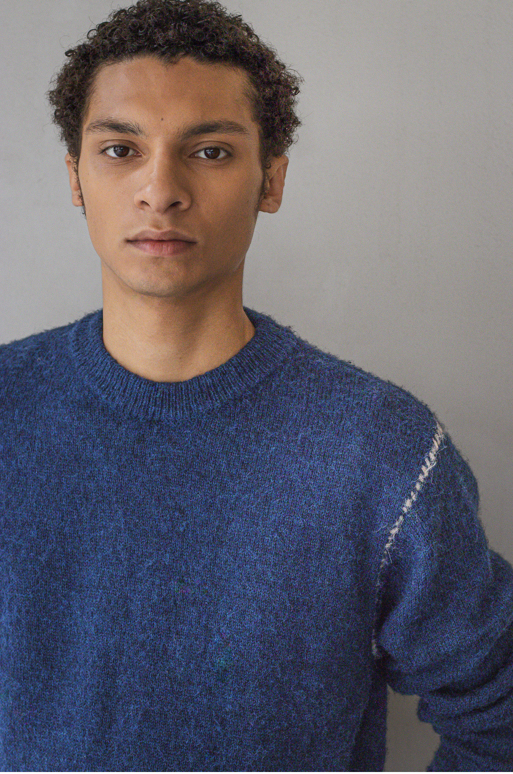 Portrait of a young man with curly hair wearing a blue sweater against a neutral background.