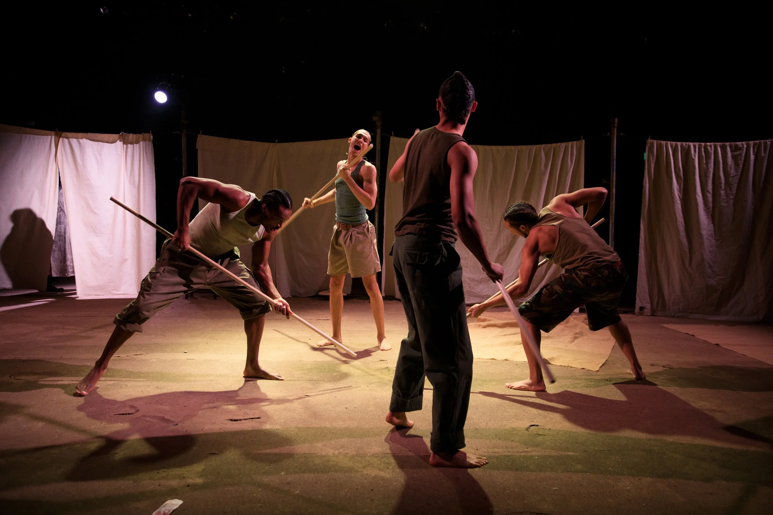 Actors performing on stage with fabric backdrops, using sticks in a theatrical scene.