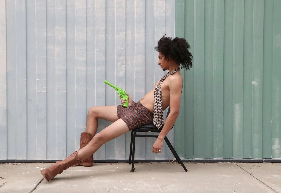 Person sitting on a chair against a colorful metal wall, holding a green water gun, wearing plaid shorts, tan boots, a tie, and no shirt.