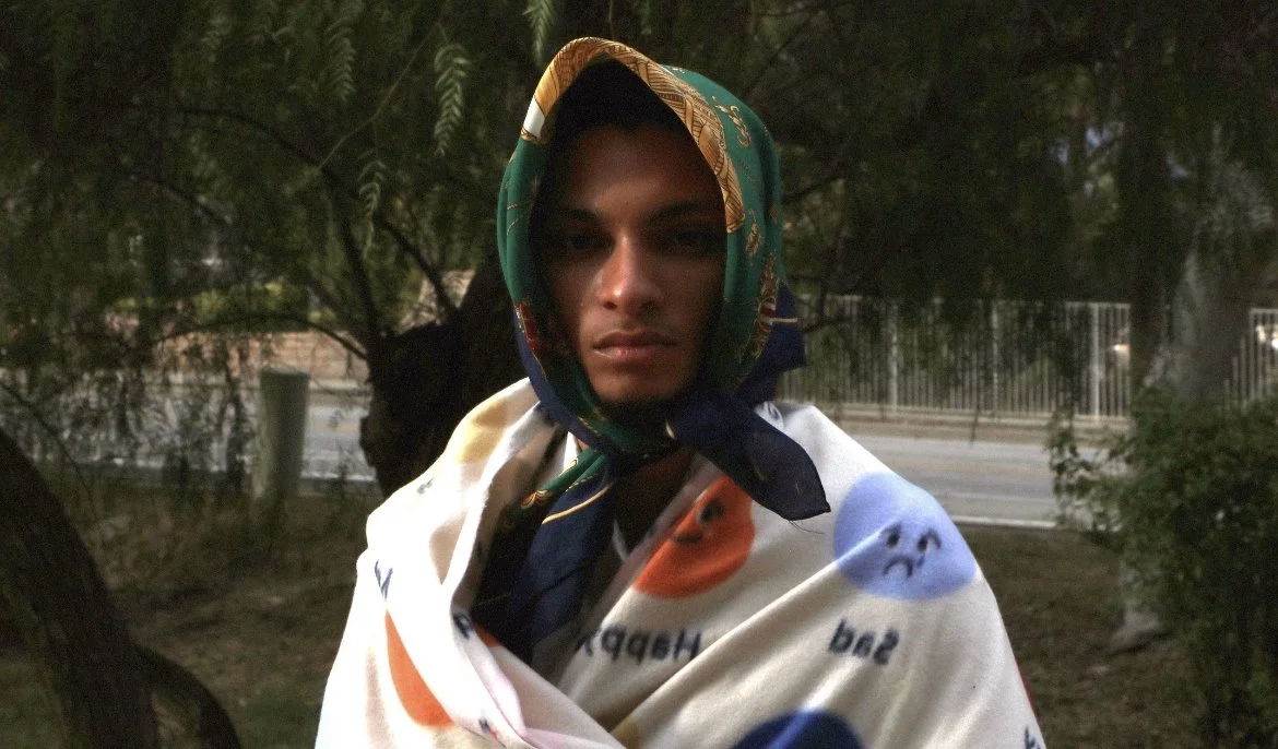 A person with a serious expression wearing a colorful scarf around their head and shoulders, standing outdoors near trees and a fence.
