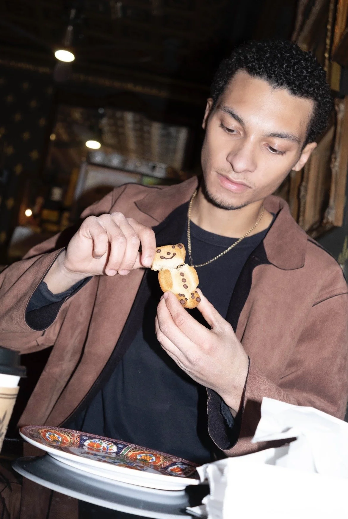 A young man with dark, curly hair and a goatee holding a gingerbread cookie shaped like a teddy bear, decorated with chocolate chips and smiling. He is wearing a brown jacket over a black shirt and is sitting at a table with colorful, decorative plat