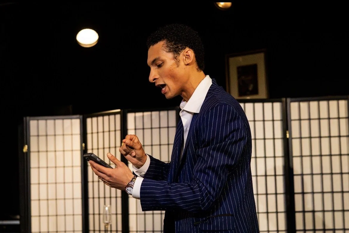 A man in a dark blue pinstripe suit looking at his phone and gesturing with his finger, standing in front of a folding screen in a dimly lit room.