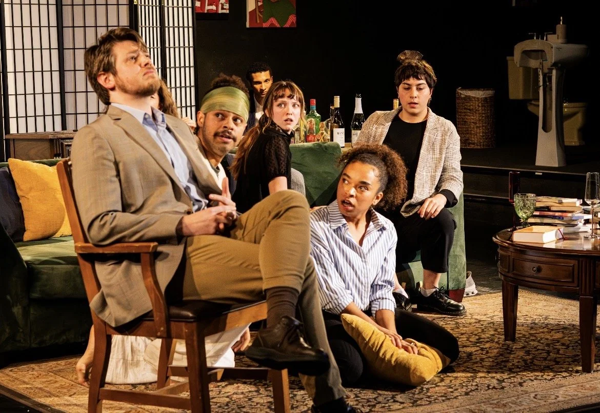 A group of six people in a living room, appearing surprised or confused, seated on a couch and floor, with a table filled with books and drinks nearby, and a bar in the background.