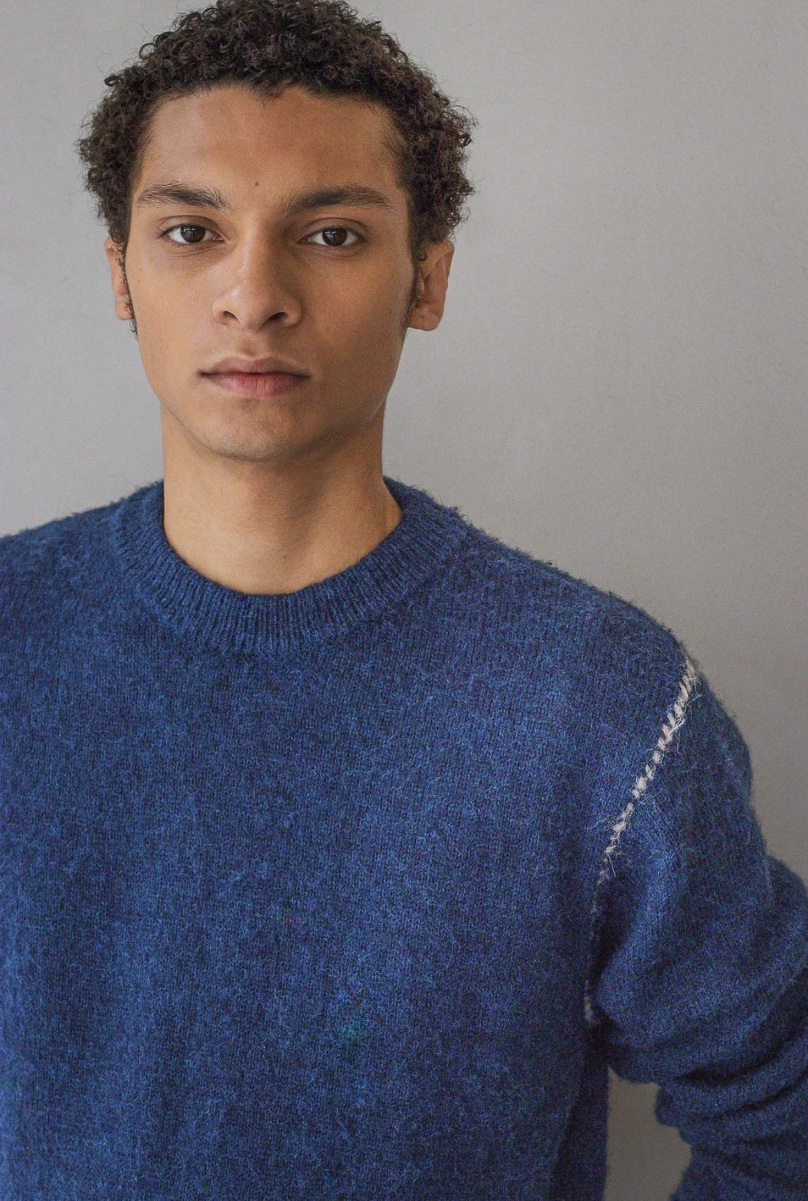 A young man with curly dark hair wearing a blue knitted sweater against a plain gray background.