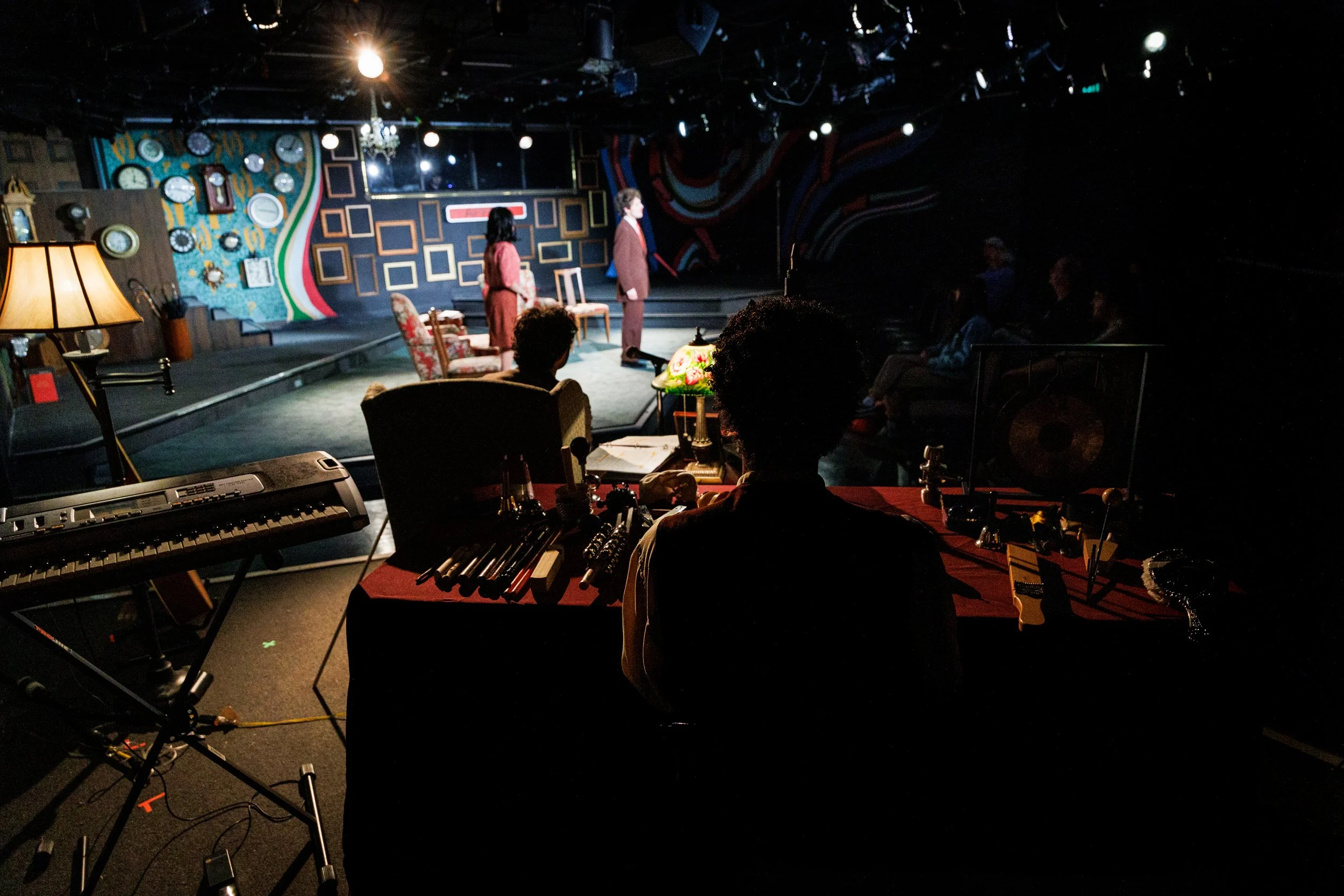 The image shows a theater stage with three actors standing and performing, with an audience seated in front. The stage is decorated with numerous clocks and a colorful abstract background. In the foreground, a sound or lighting technician is working 