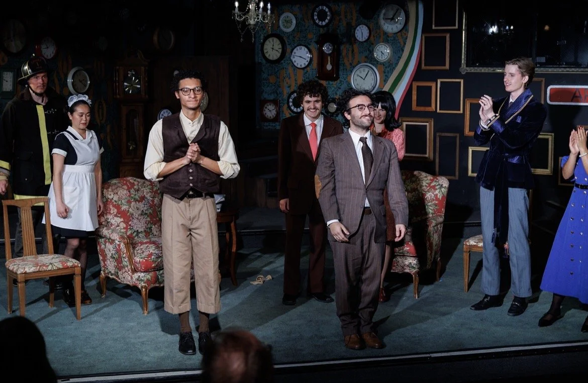 The stage of a theatrical play with actors in costumes, some in formal attire, standing and clapping. The background has many clocks on the wall and a chandelier hanging from the ceiling.
