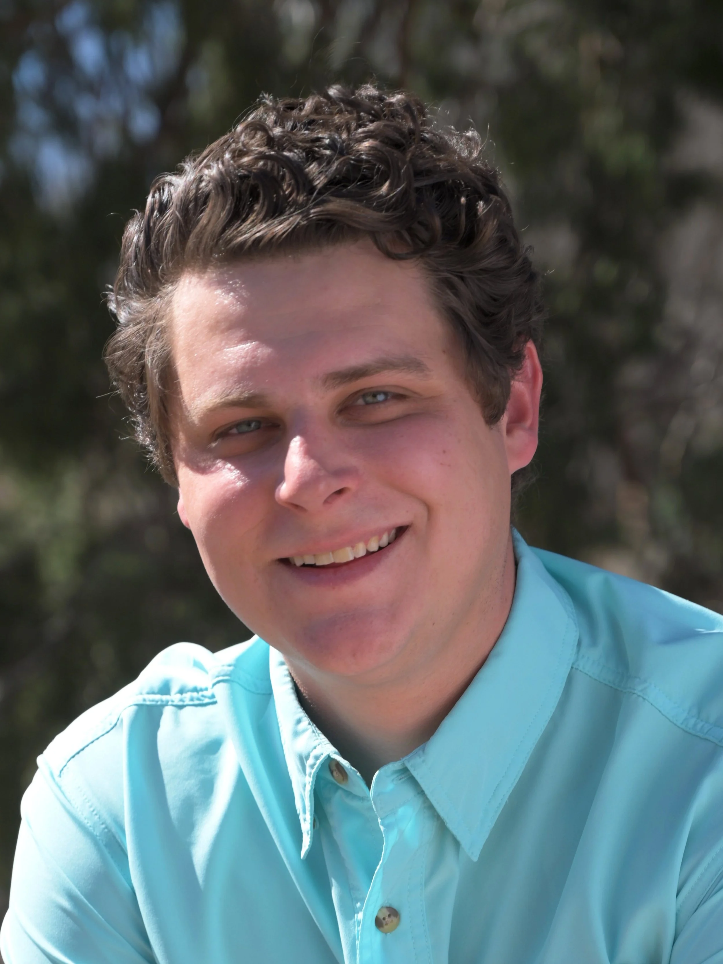 Close-up of a young man smiling outdoors, wearing a light blue button-up shirt, with trees in the background.