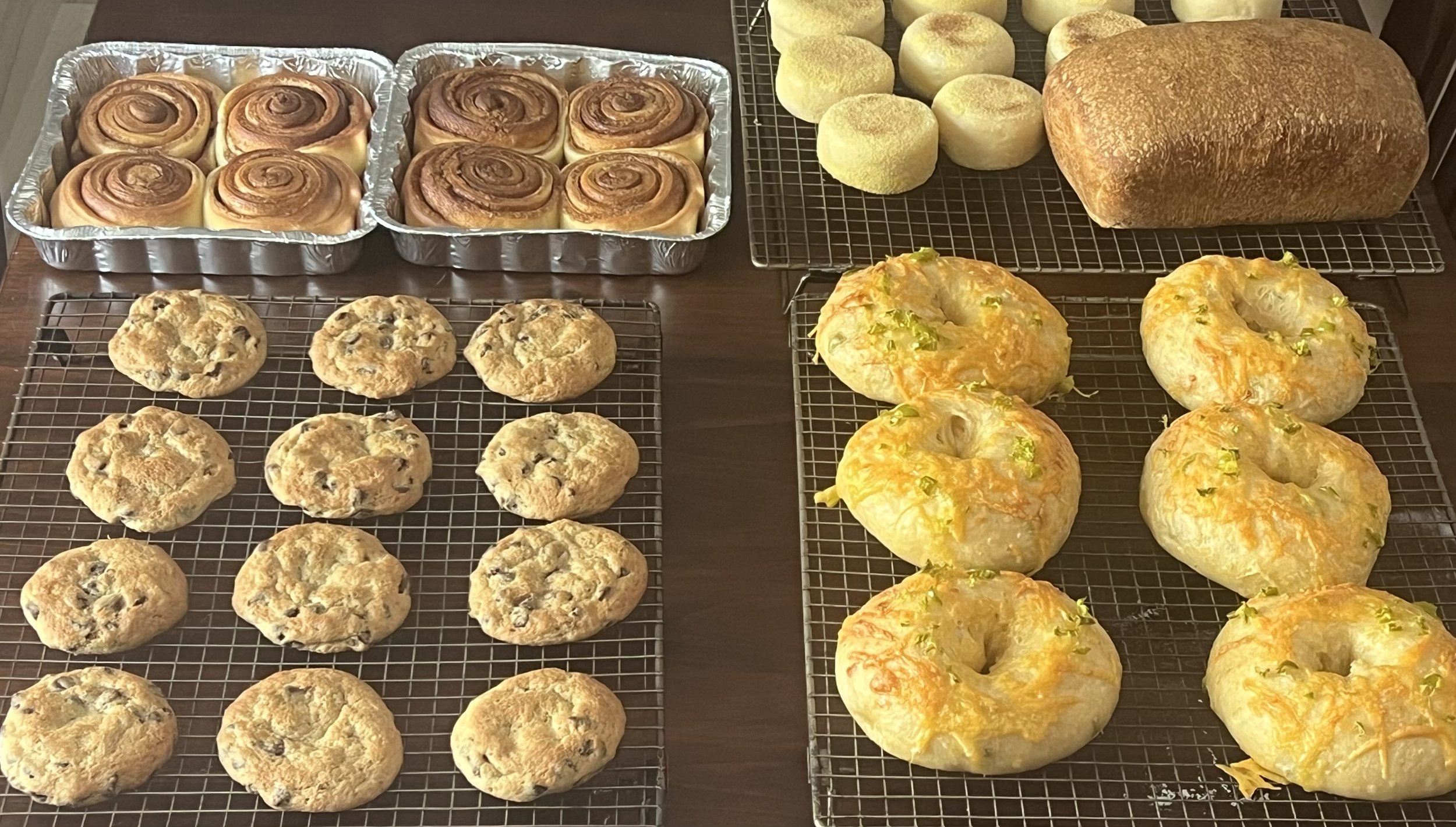 Assorted baked goods including cinnamon rolls, cookies, hamburger buns, and bagels cooling on wire racks and trays.
