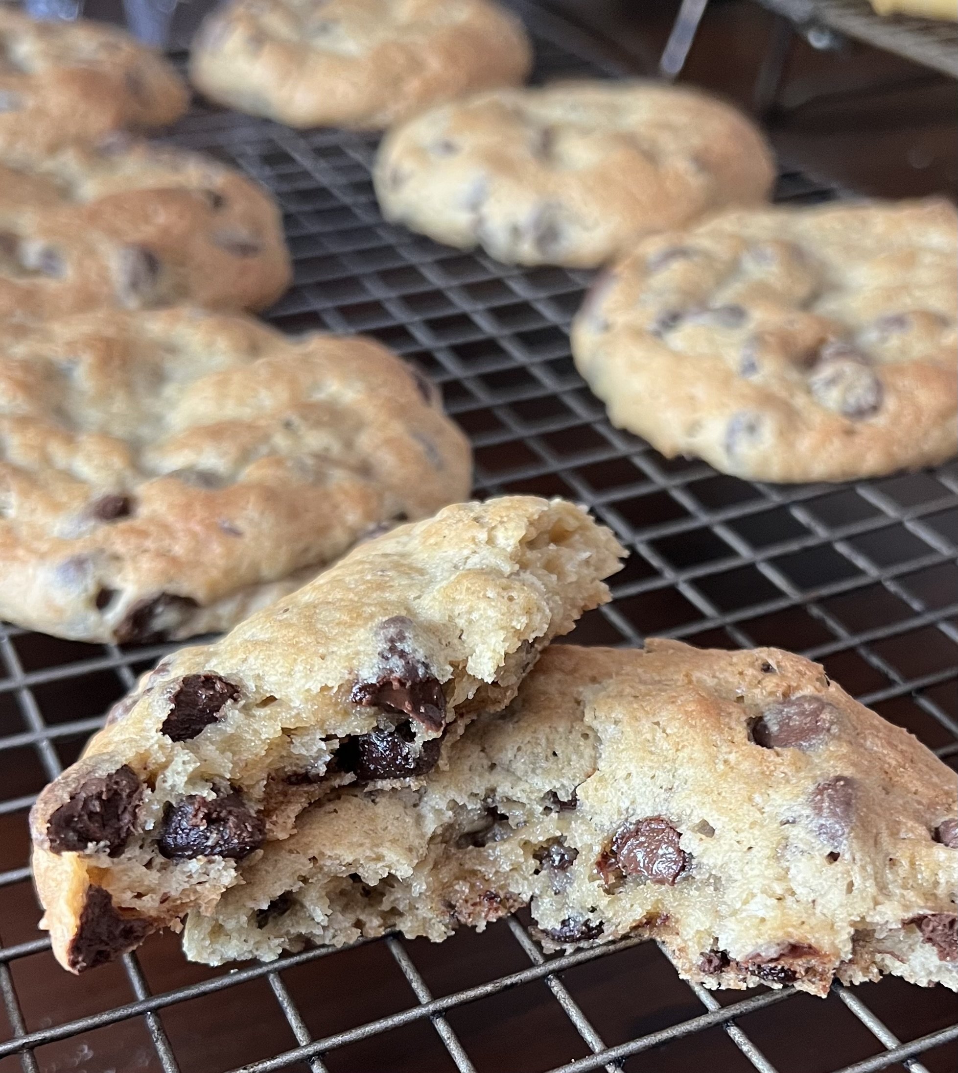 Close-up of a chocolate chip cookie broken in half, showing the inside with melted chocolate chips. Several whole cookies are cooling on a wire rack in the background.
