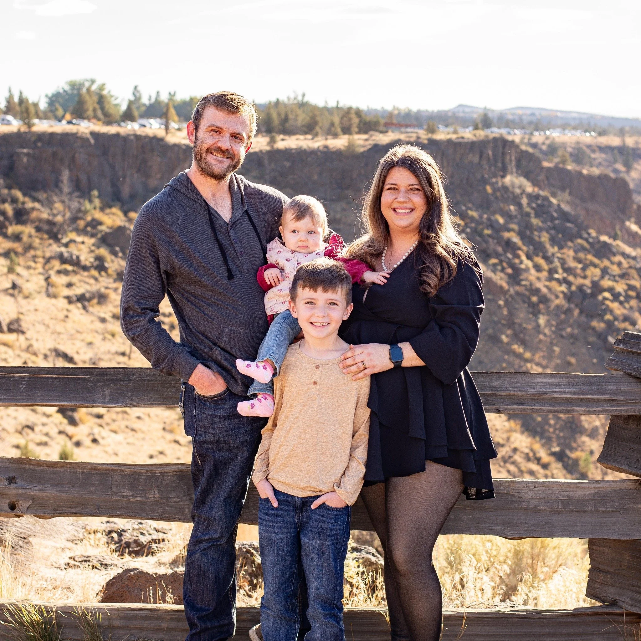A family of four outdoors with a scenic canyon background, including a bearded father, smiling mother, a young boy, and a baby girl, all smiling at the camera.