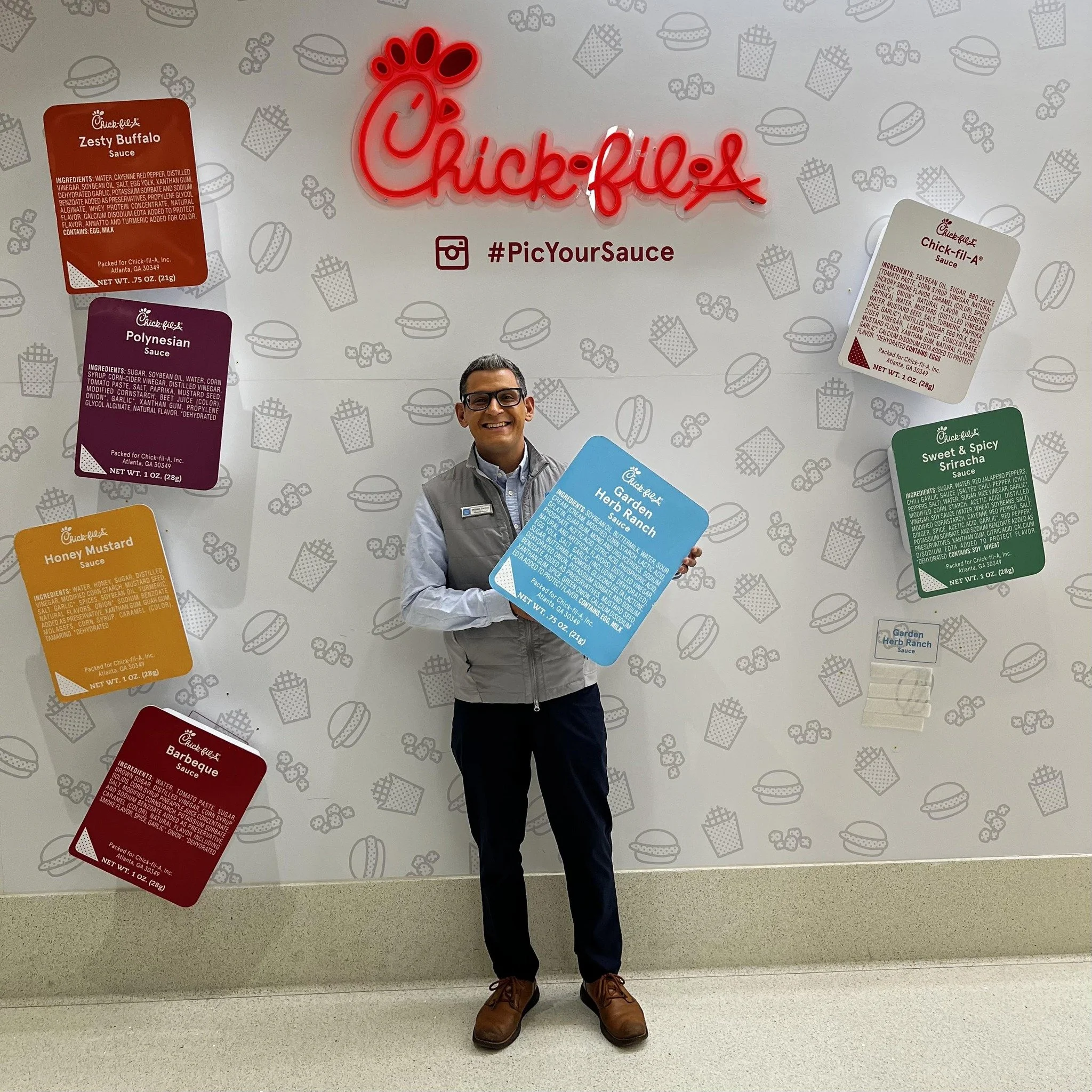 A man in business casual attire, wearing glasses and smiling, standing in front of a wall with Chick-fil-A branding and menu cards. He is holding a large blue card titled 'Garden Herb Ranch,' with other colorful cards for different Chick-fil-A sauces displayed on the wall behind him.