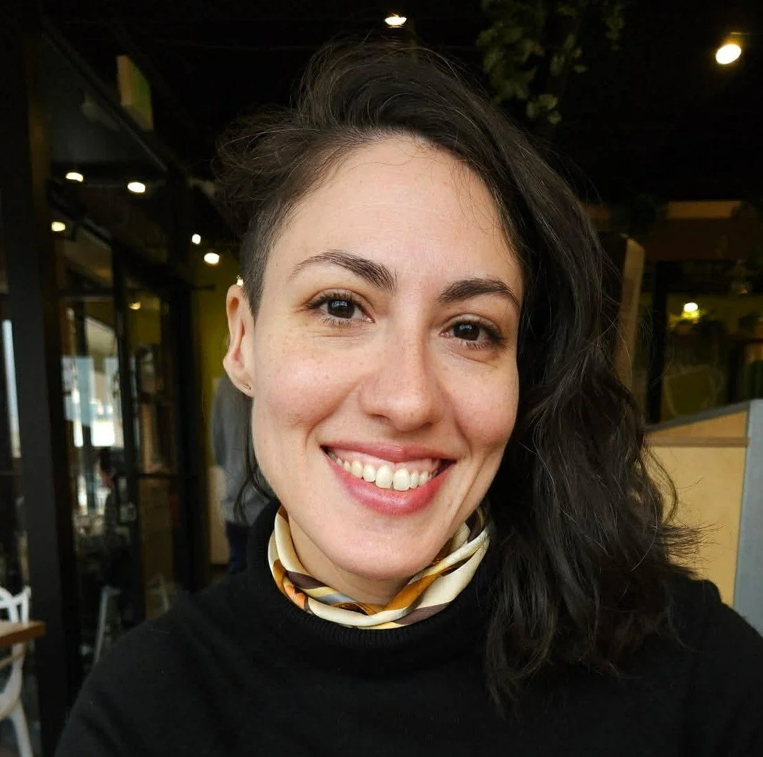 Close-up of a smiling woman with black hair and a scarf, indoors at a cafe or restaurant.