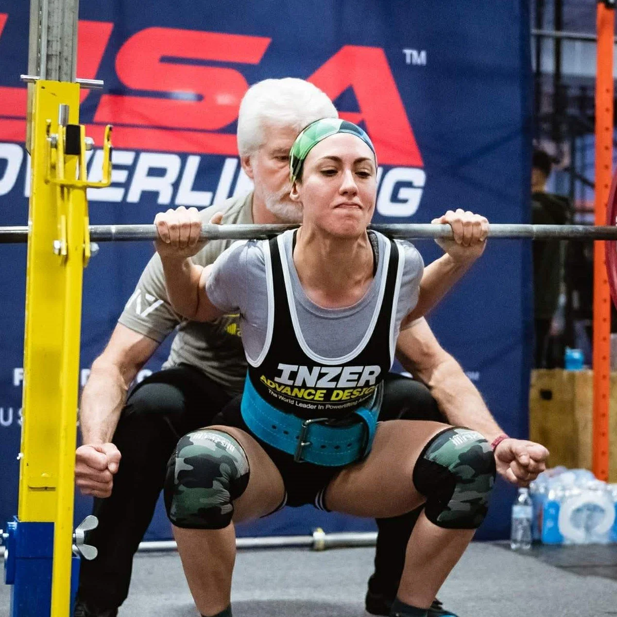 Woman doing squat with a barbell at a powerlifting competition, with a man standing behind her guiding her, in an indoor gym setting.