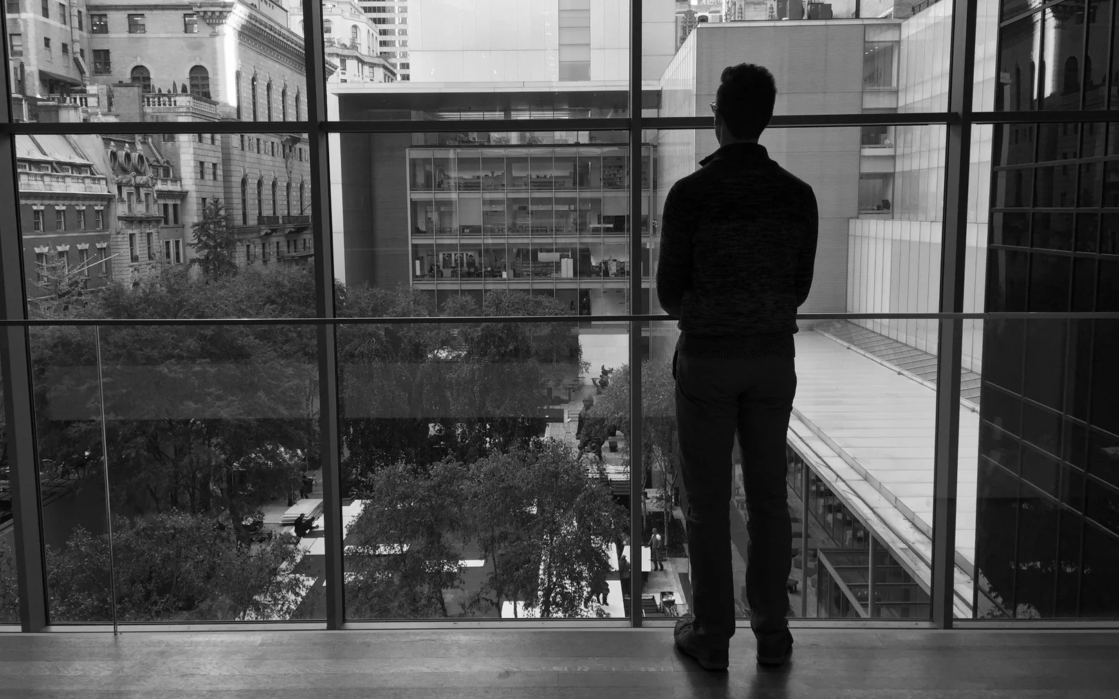 A man stands looking out a large window at a city street below during daytime. The city features tall buildings and trees.