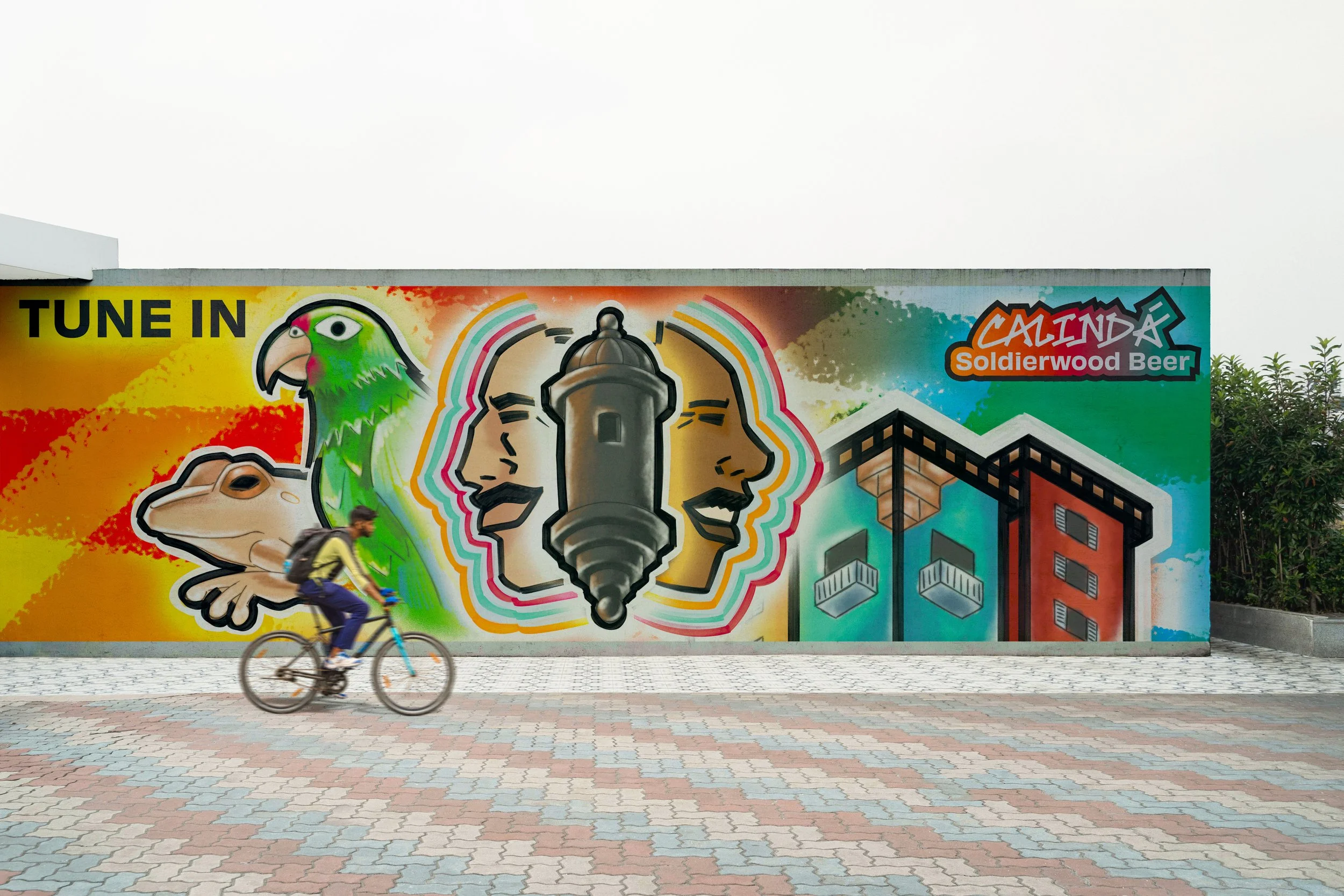 A picture of a painted mural depicting a stone watchtower, stylized profiles of a man and woman, a parrot, a frog, and two brightly colored buildings on a colorful swirly background. The mural reads "Tune In" and "Calindá: Soldierwood Beer" on the le